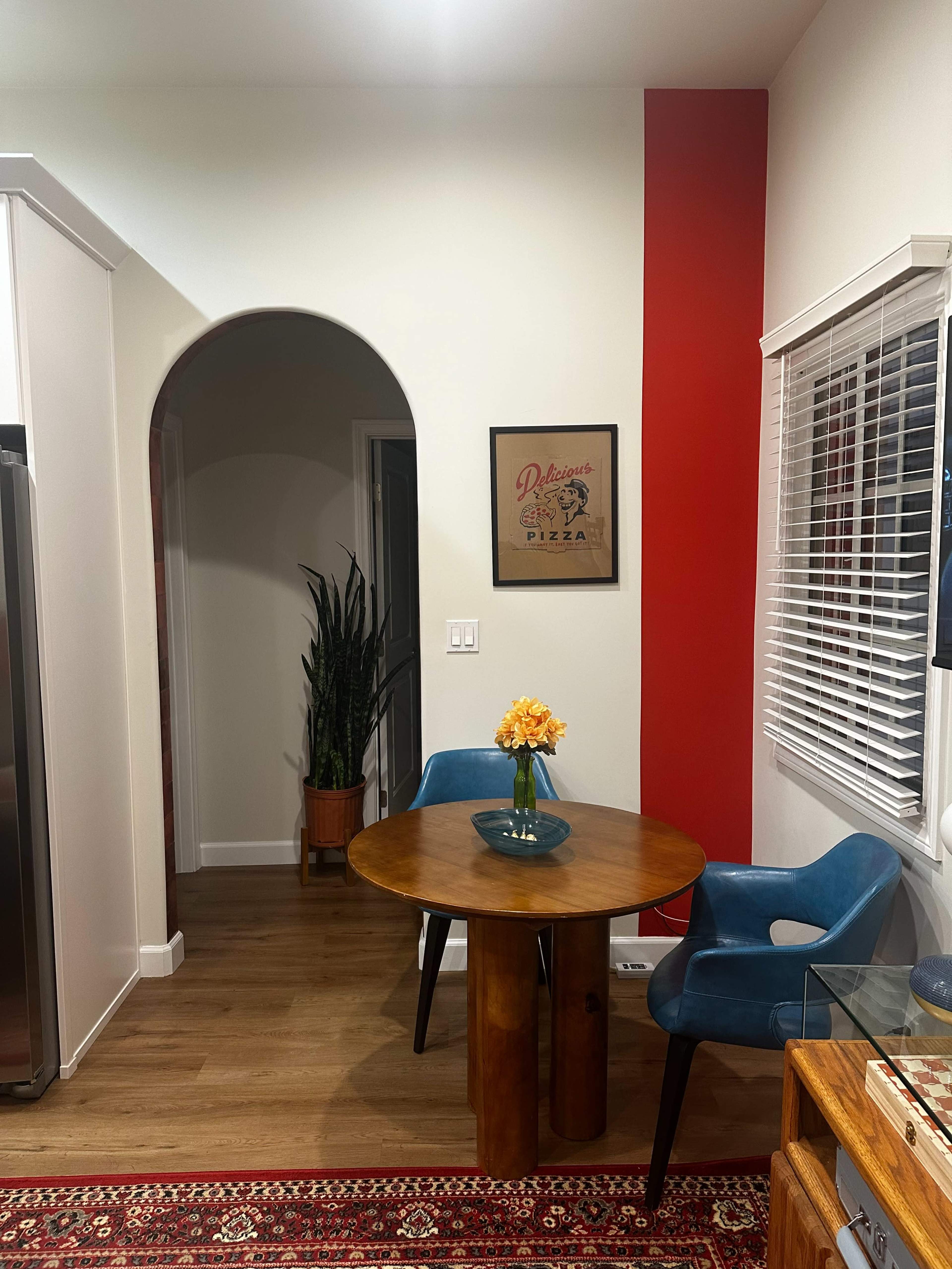 The image shows a small dining area with a round wooden table, two blue chairs, a plant in a pot, and a wall featuring a red accent with a framed pizza poster.
