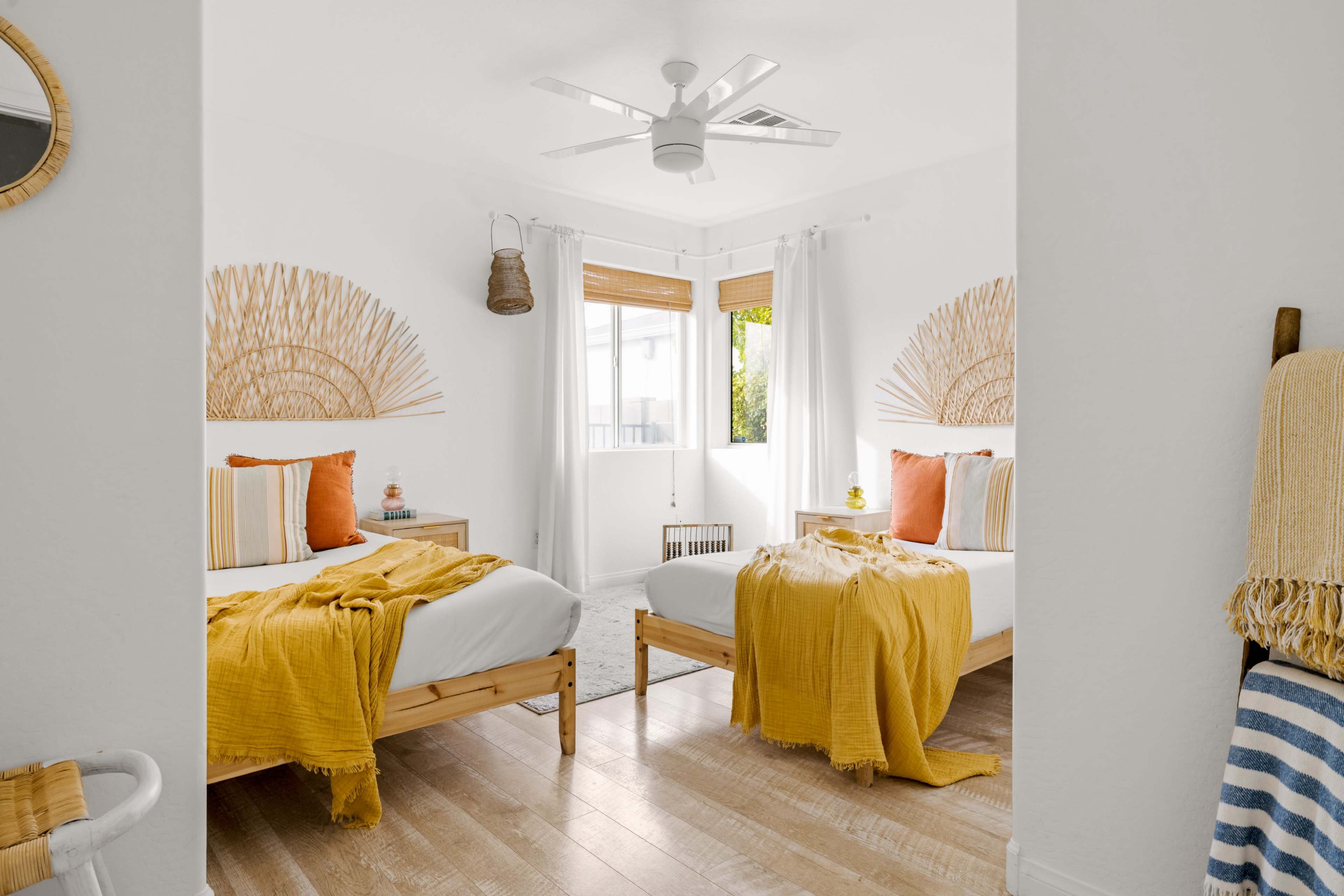 The image shows a bright and airy bedroom featuring two twin beds with yellow throws, decorative wall accents, and large windows providing natural light.