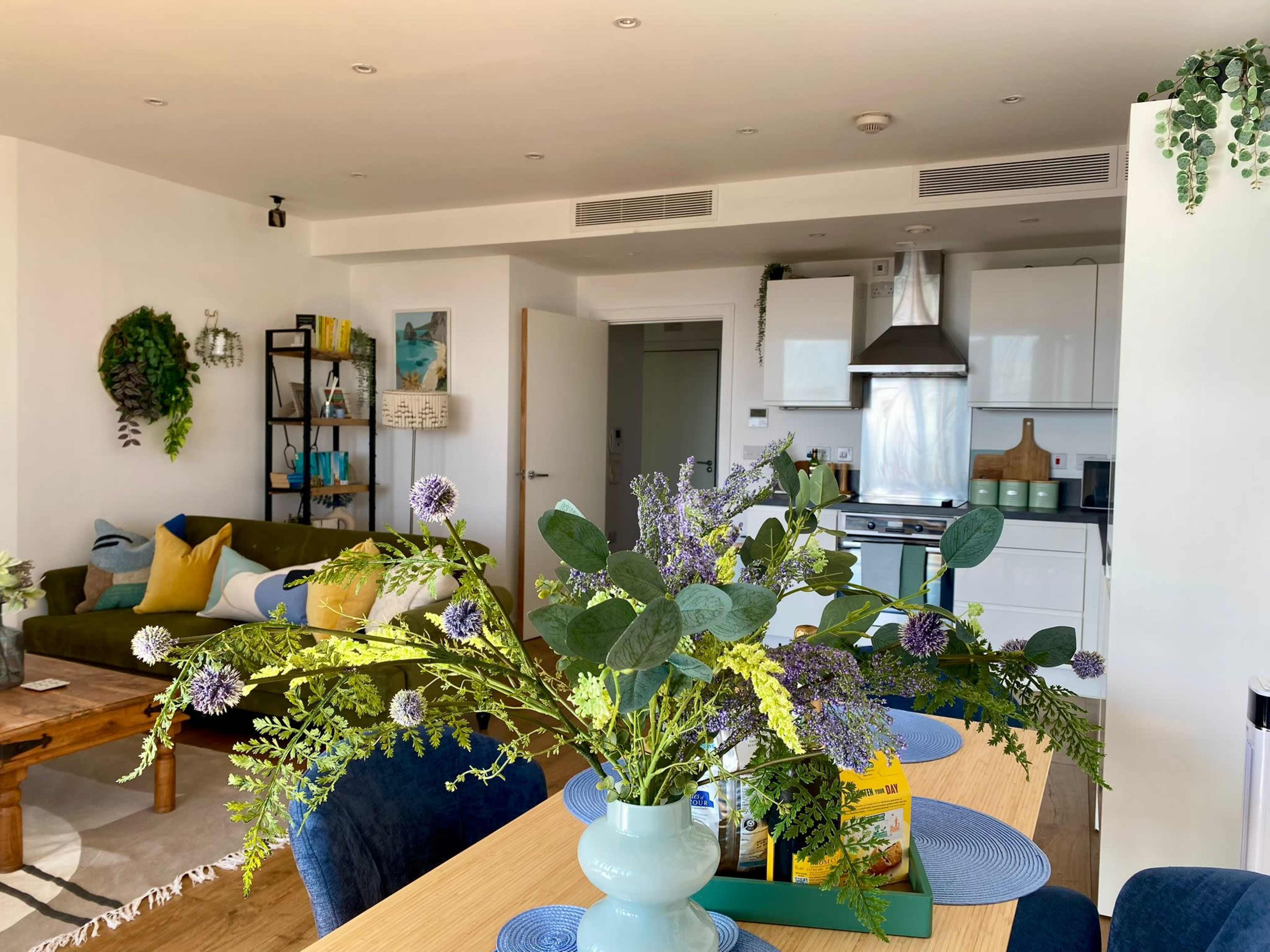 A modern apartment showcases a dining area with a floral centerpiece, surrounded by blue chairs, and an open kitchen visible in the background.