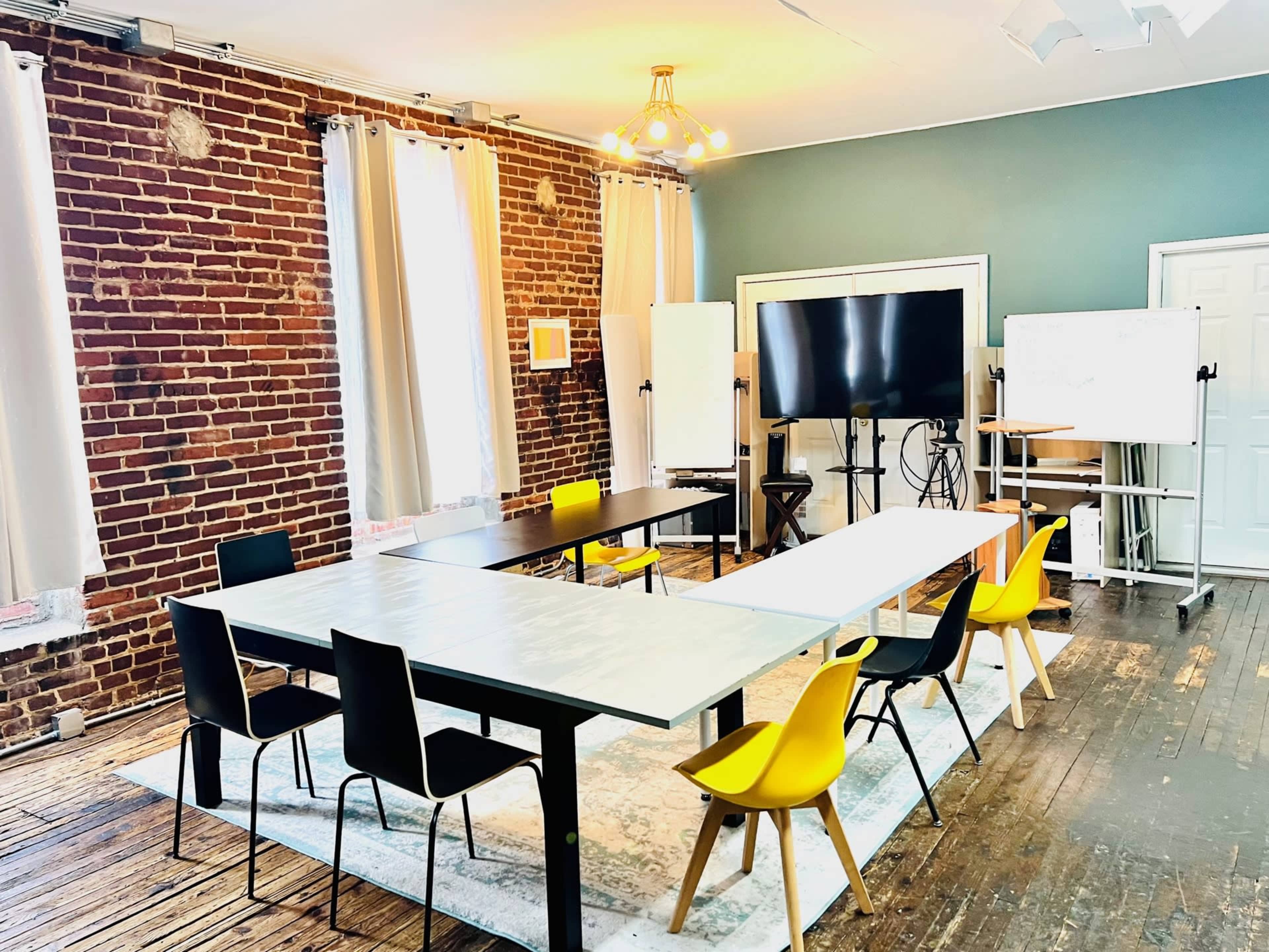 A bright meeting room features multiple tables arranged in a U-shape, with black and yellow chairs, brick walls, and large windows allowing natural light.