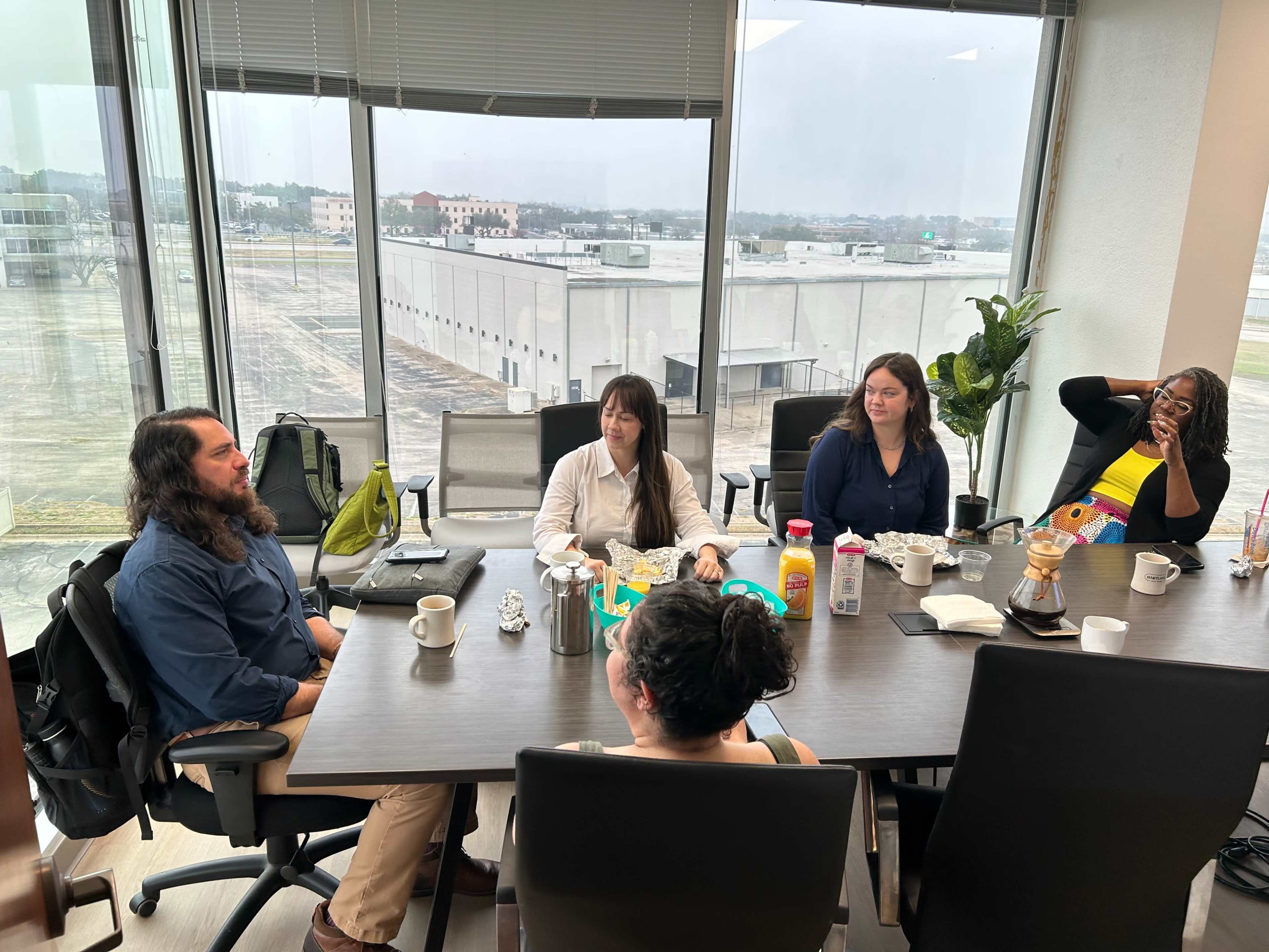 A group of five people is seated around a conference table with various beverages and snacks, while large windows display an industrial landscape outside.
