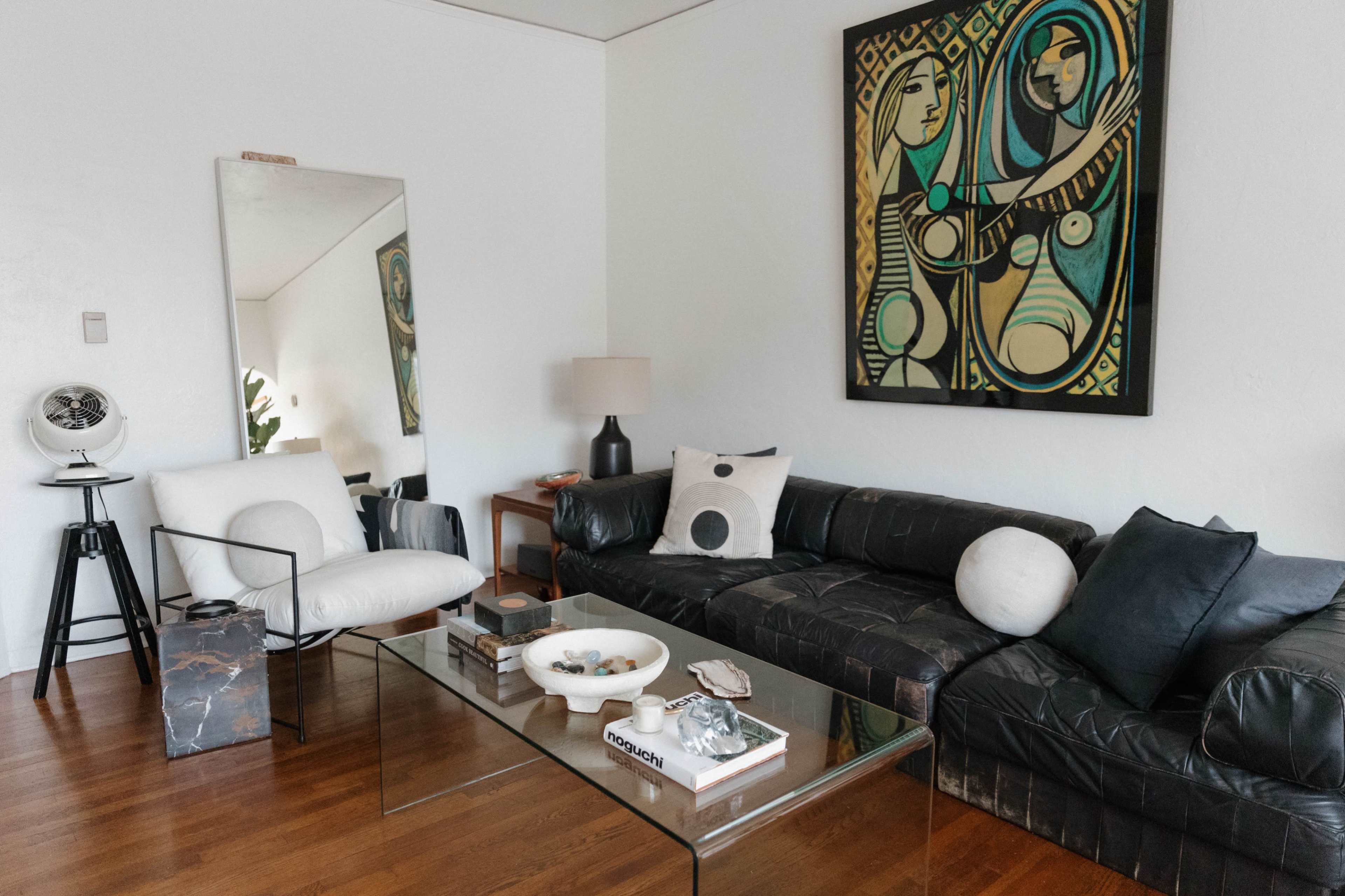 A modern living room featuring a black leather sectional sofa, a glass coffee table, a large mirror, and a vibrant abstract painting on the wall.
