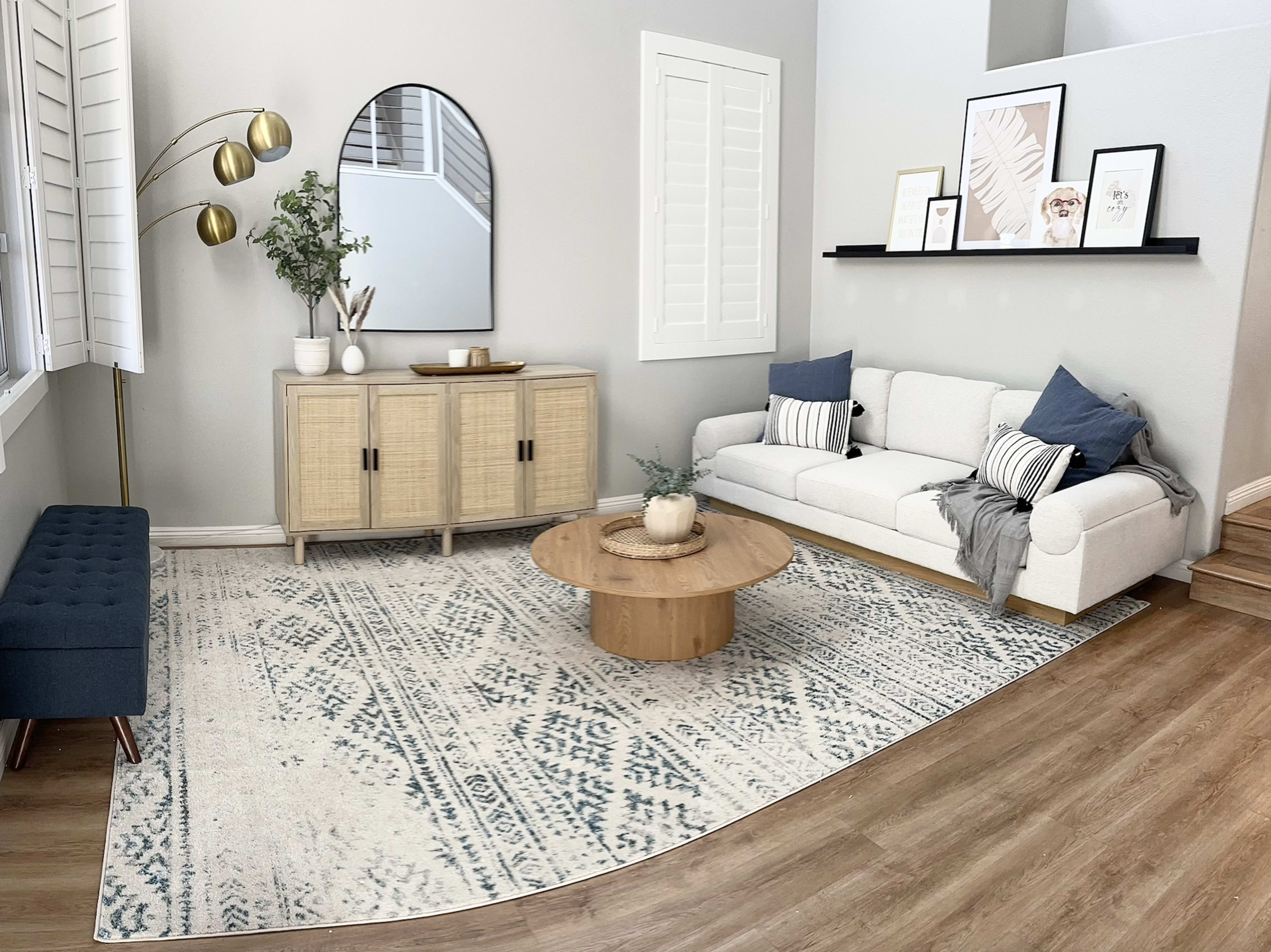 A modern living room features a white sofa, a round wooden coffee table, and an area rug with a geometric pattern, accompanied by a sideboard and decorative plants.