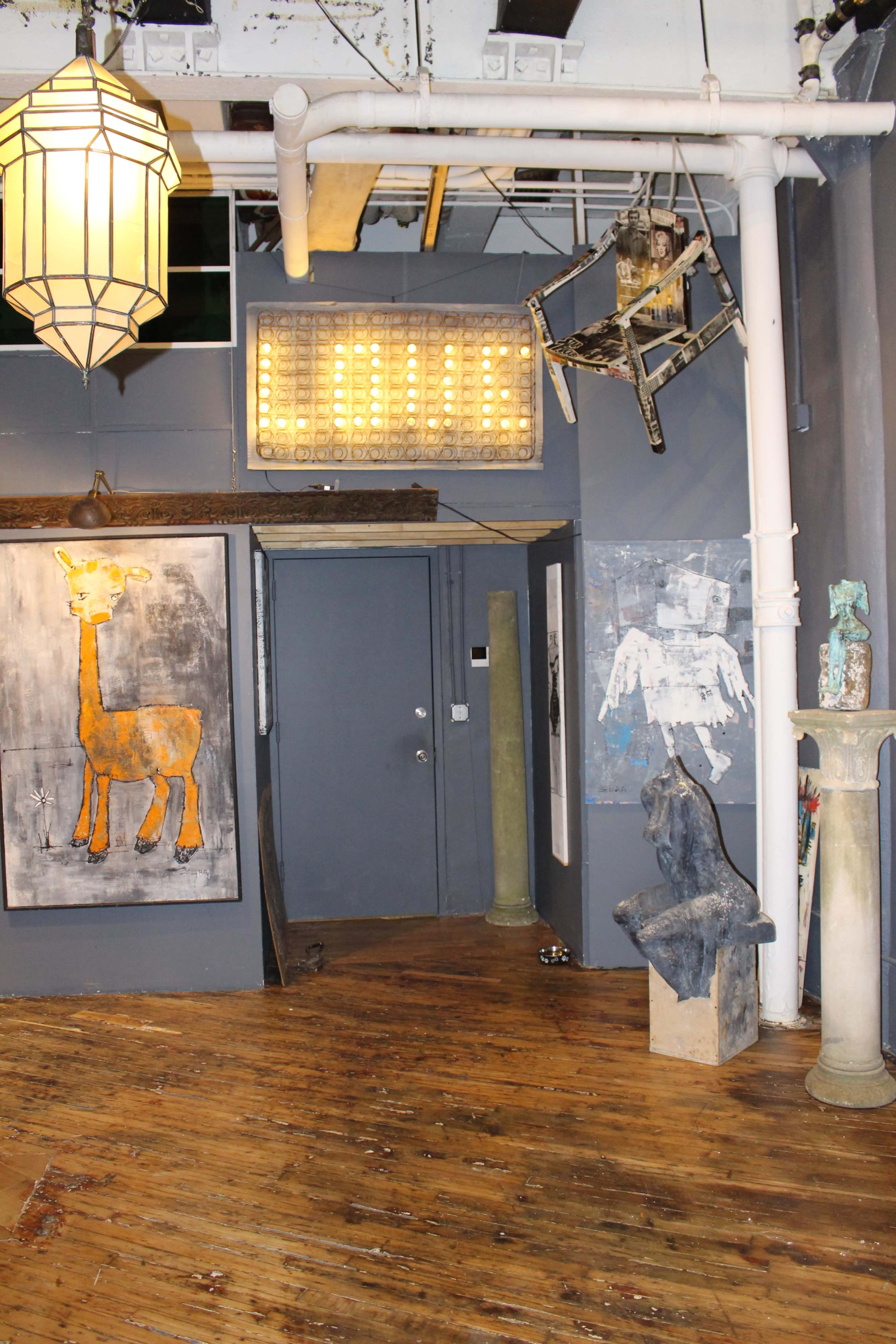 The image shows an art gallery interior featuring a giraffe painting, sculptures, a hanging chair, and a vintage light fixture in a wooden-floored space.