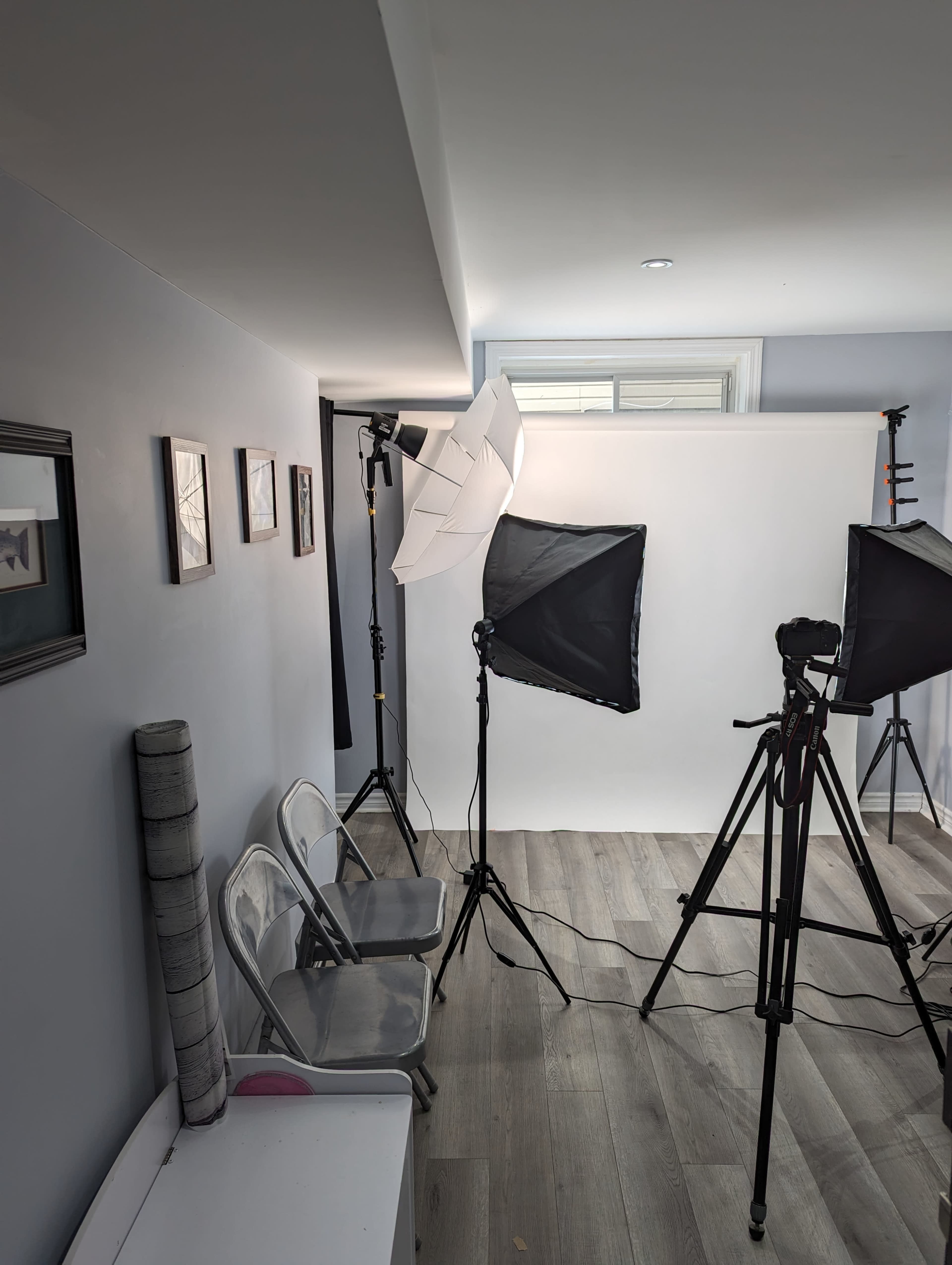 A photography studio setup with two light umbrellas, tripods, and empty chairs against a gray wall.