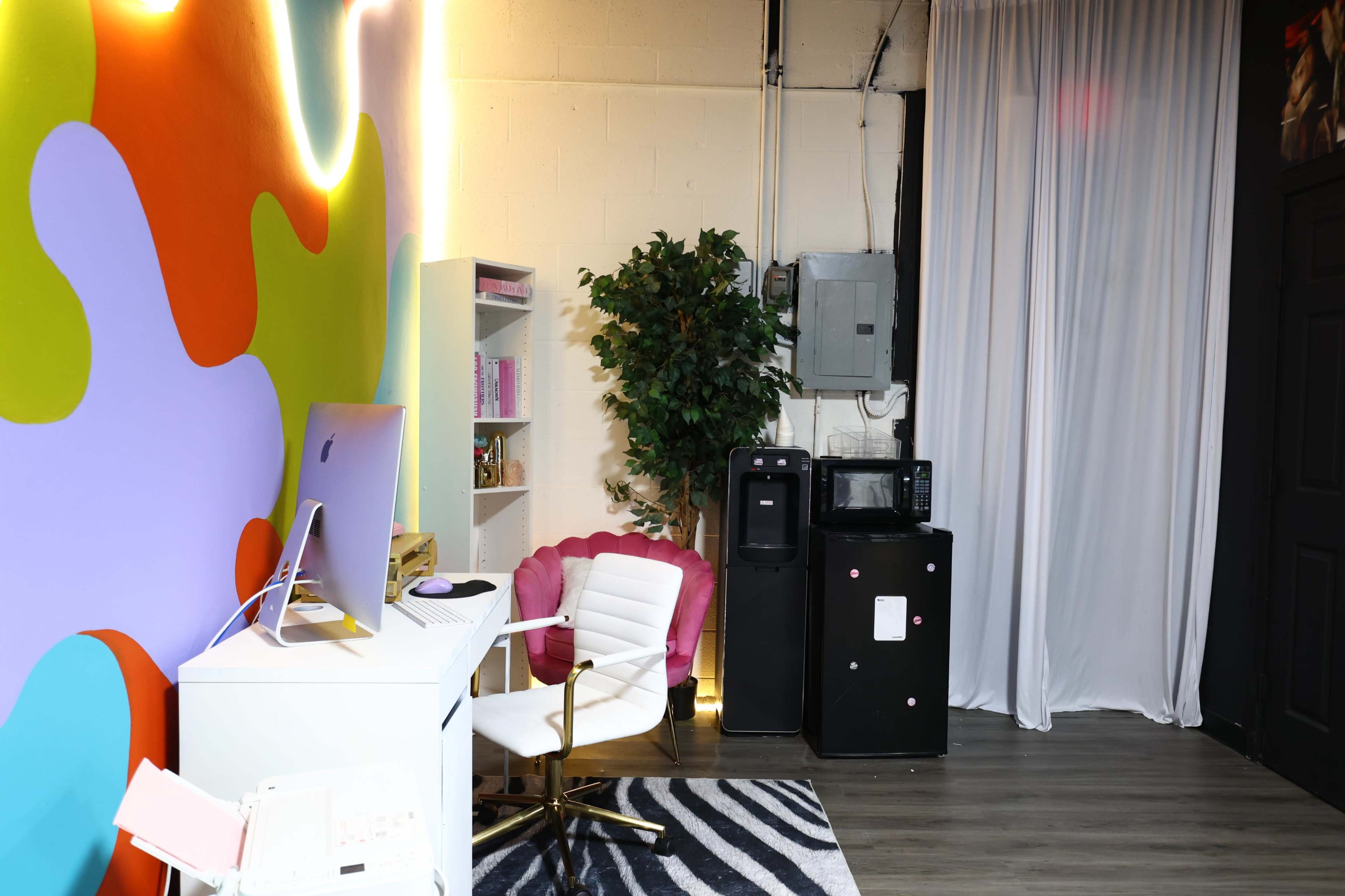 The image shows a colorful wall with a white office desk and chair, a plant, and a black refrigerator next to a microwave in a room with gray flooring.