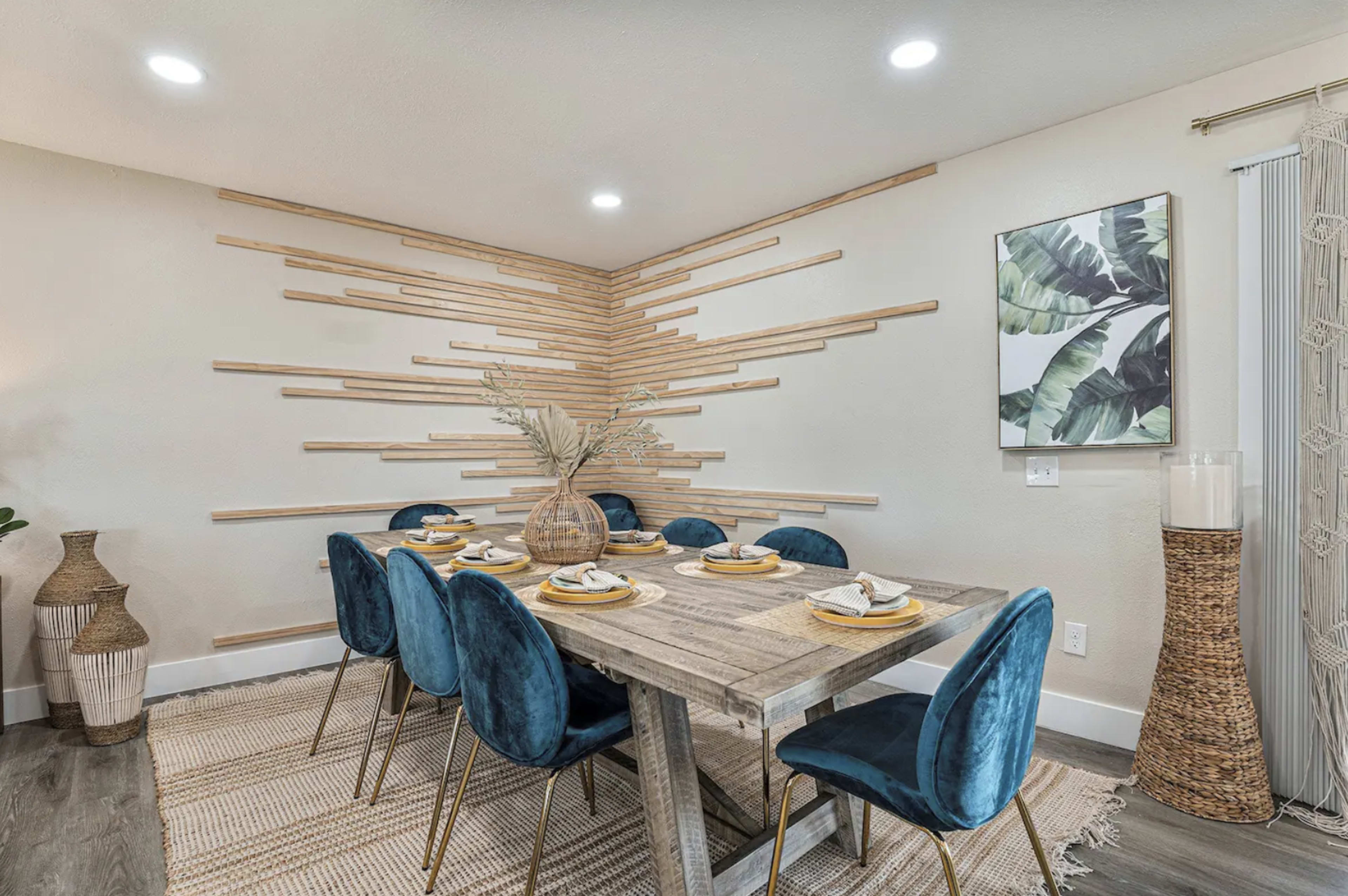 A dining area features a wooden table set for eight with blue chairs, against a wall adorned with wooden strips and a tropical-themed artwork.