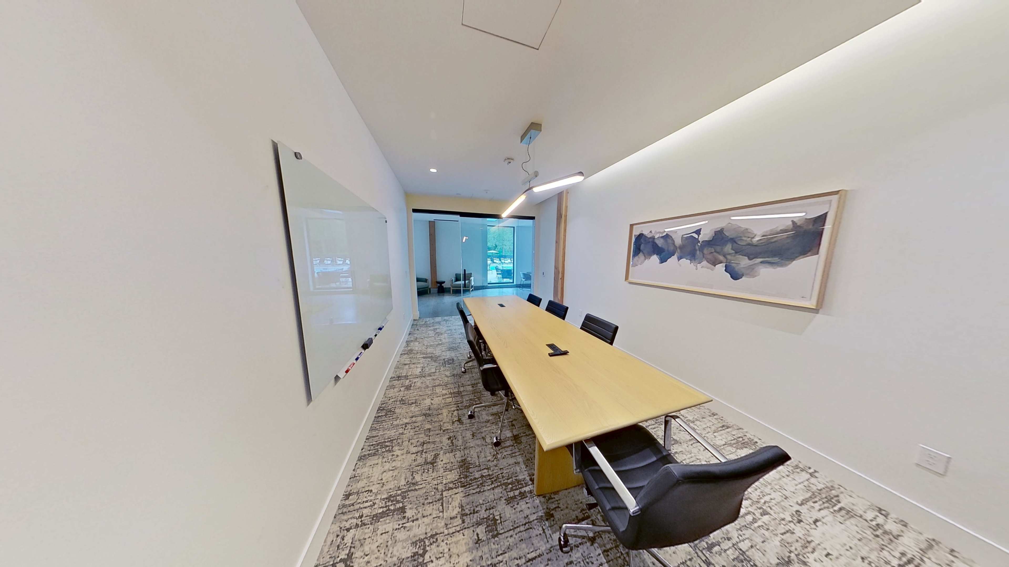 A modern conference room features a long wooden table surrounded by black chairs, with a whiteboard on one wall and a large artwork on another.