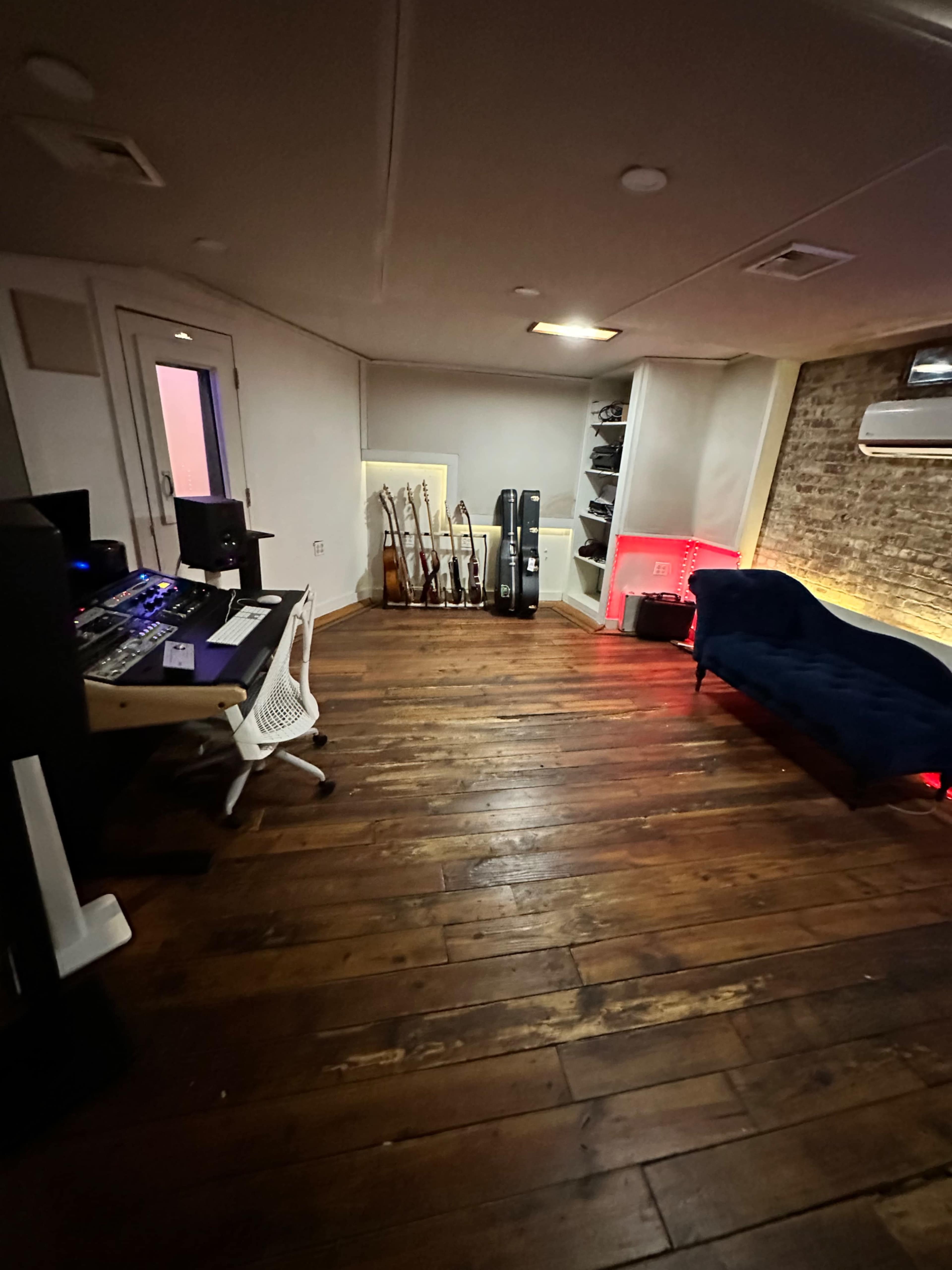 The image shows a music studio with wooden floors, a control desk featuring equipment, and a couch beside a display of guitars in a corner.