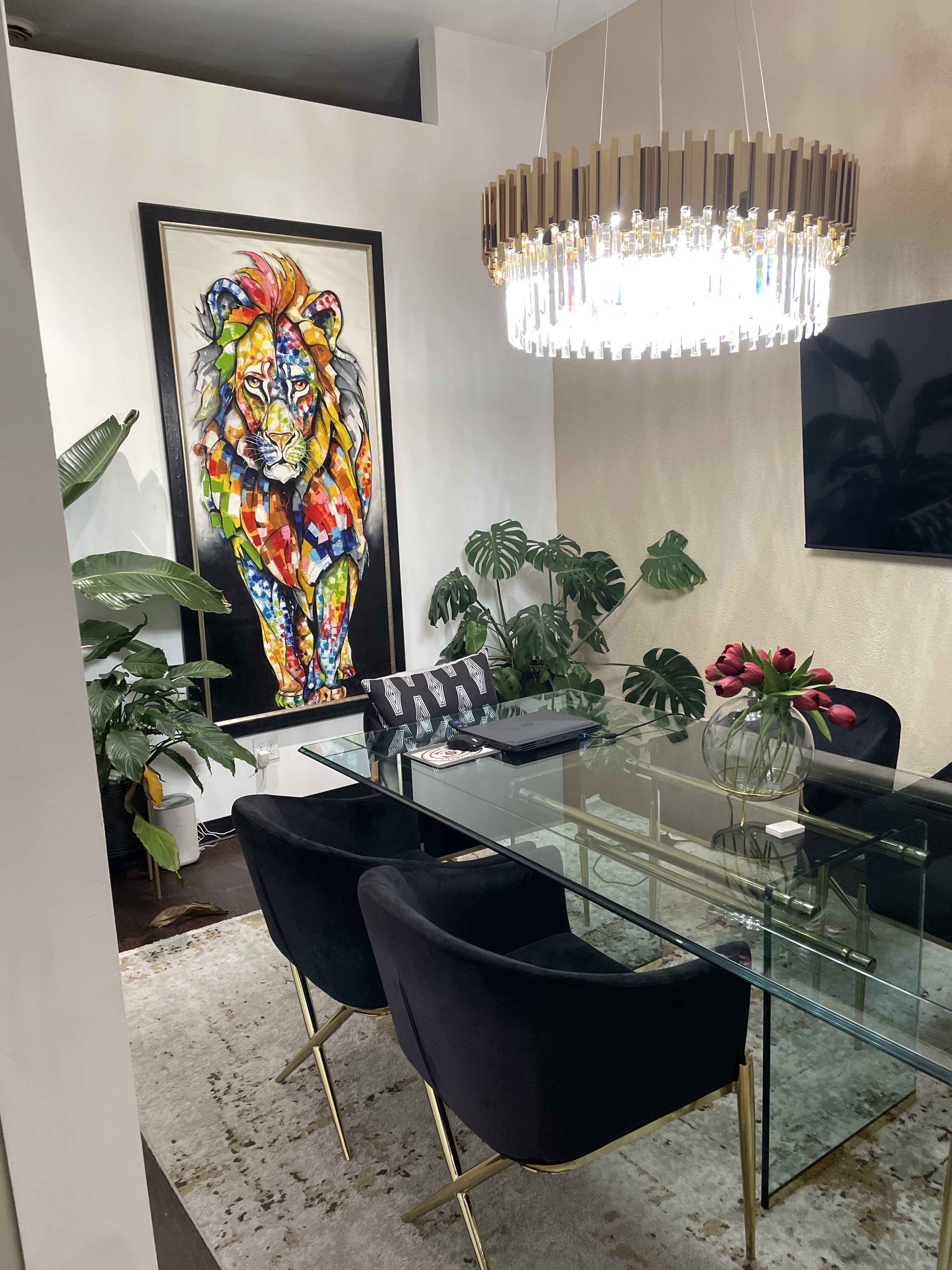A stylish dining area featuring a glass table surrounded by black velvet chairs, a colorful lion painting on the wall, and a large chandelier overhead.