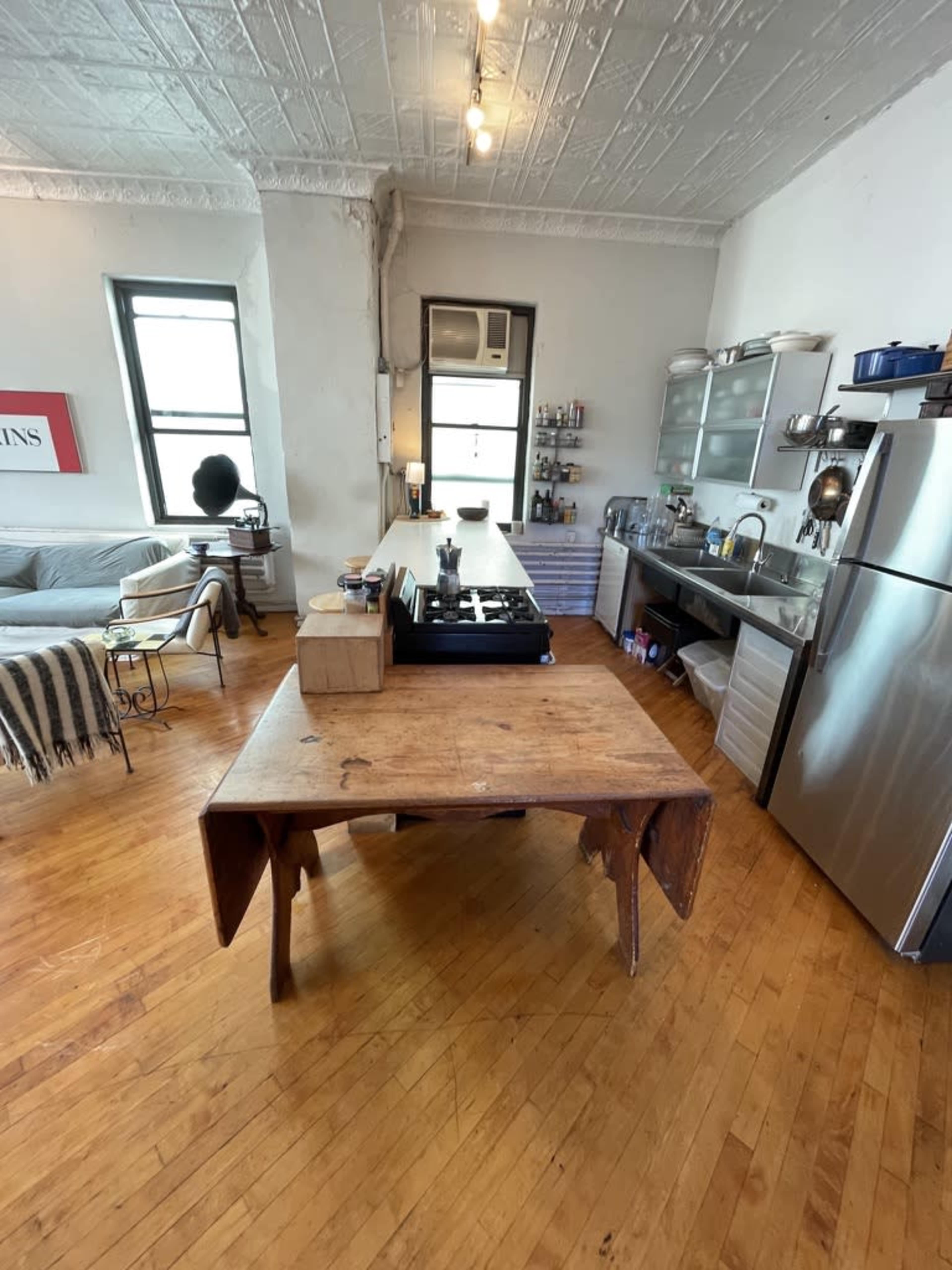 A bright kitchen features a central wooden table with a gas stove on top, surrounded by stainless steel appliances and shelves with various kitchen items.