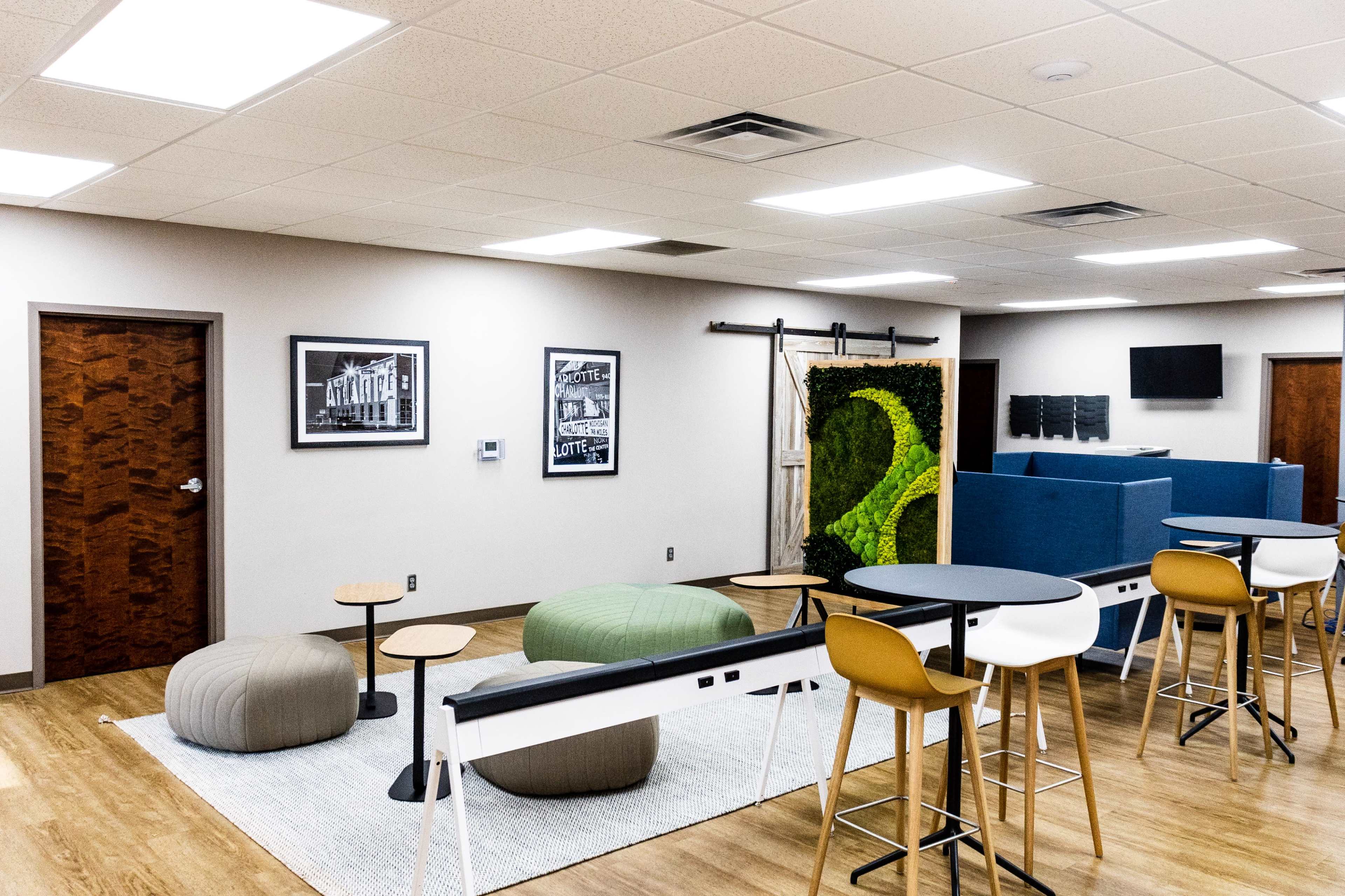 The image shows a modern office lounge area with circular seating arrangements, a decorative green wall, and a mix of tables and high chairs.