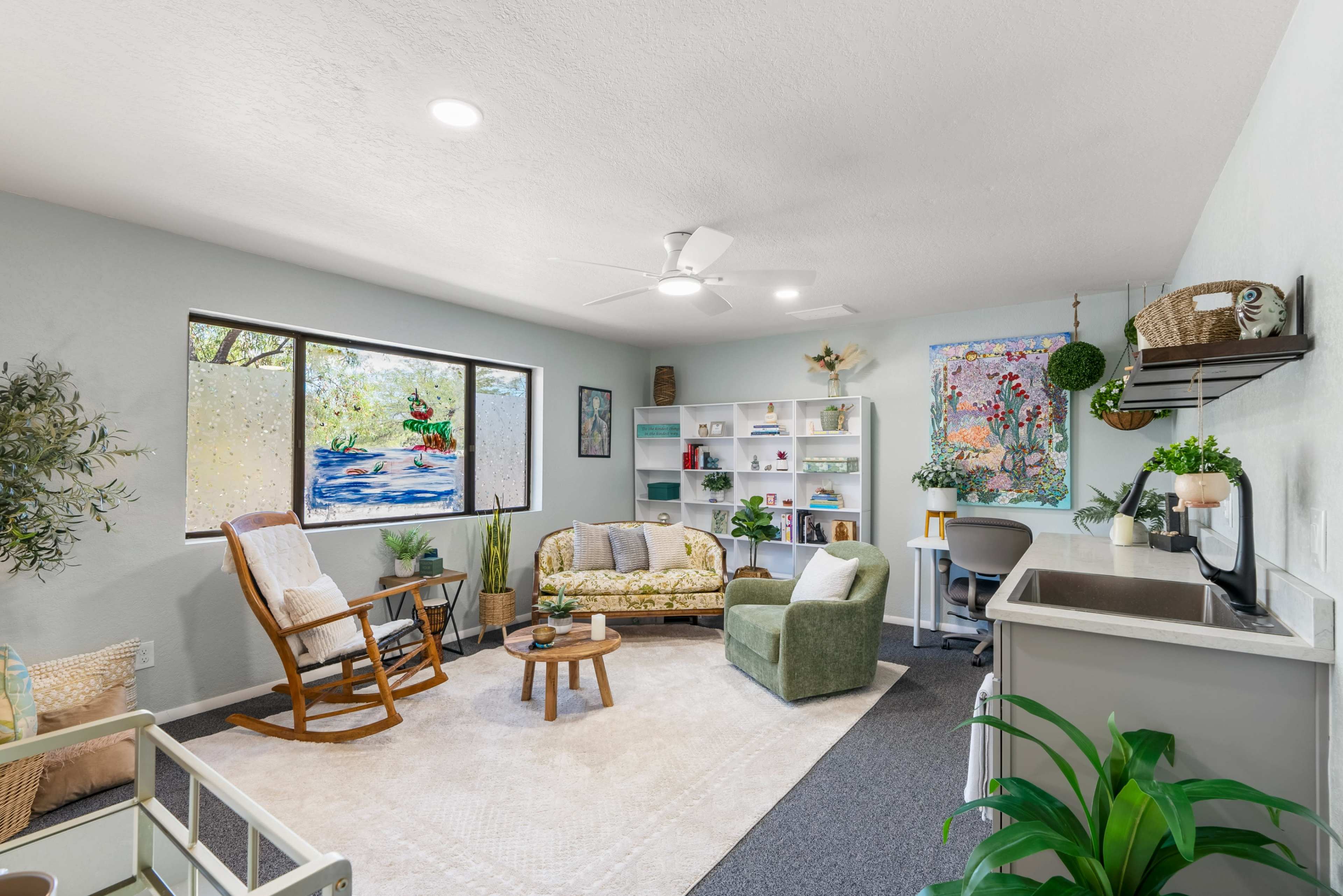 The image shows a modern living room with a green couch, a rocking chair, a small coffee table, and colorful wall art near a large window.
