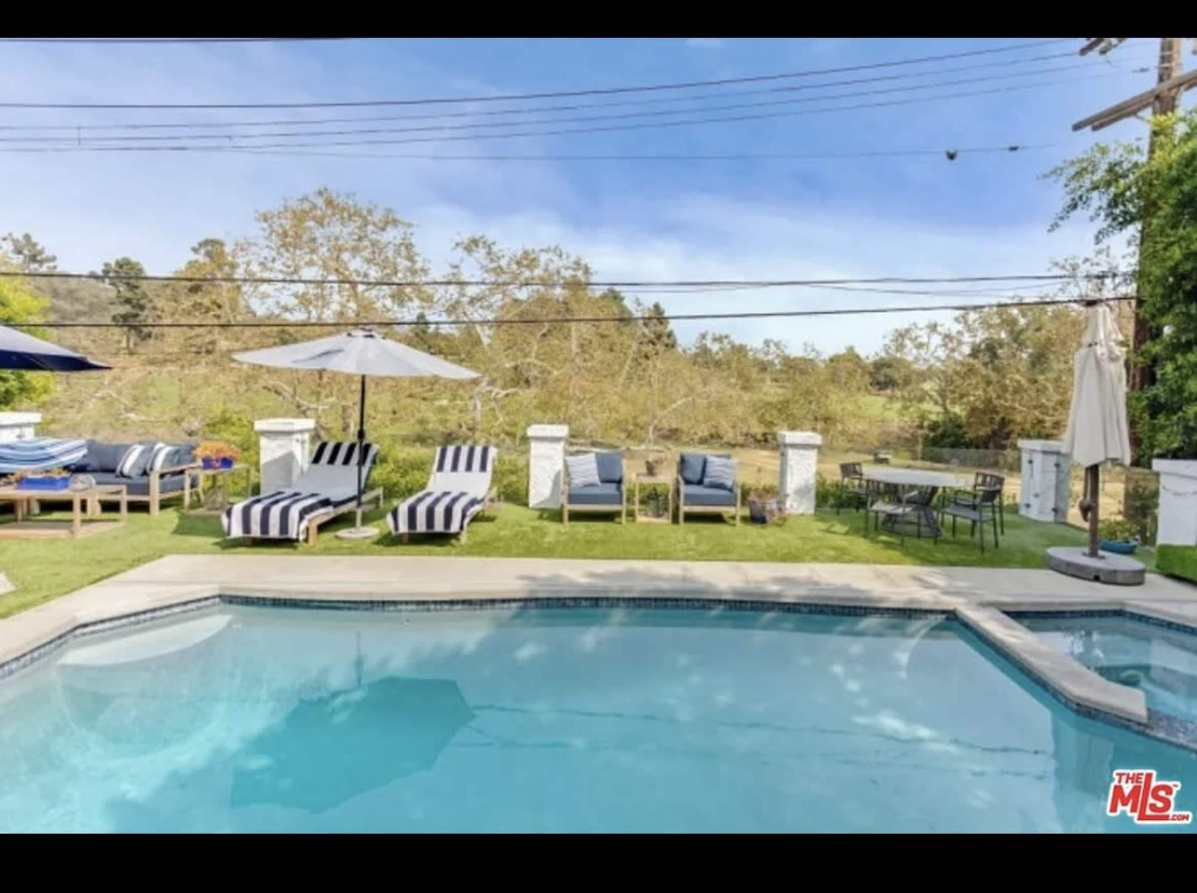 A swimming pool is surrounded by lounge chairs and umbrellas, with a grassy area and trees in the background.