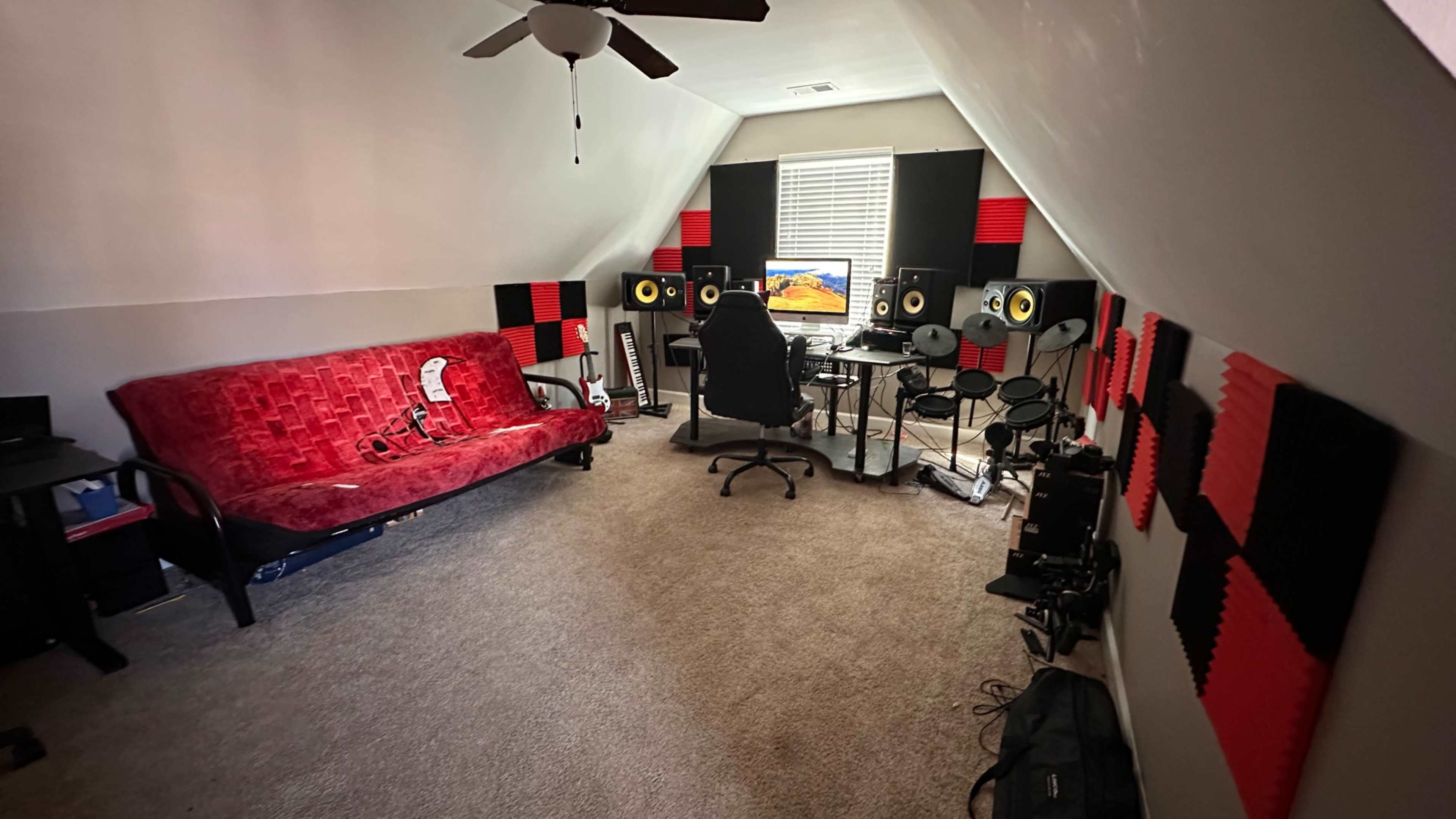 The image shows a music production studio in an attic with a red and black color scheme, featuring a desk with monitors, speakers, and soundproofing panels.