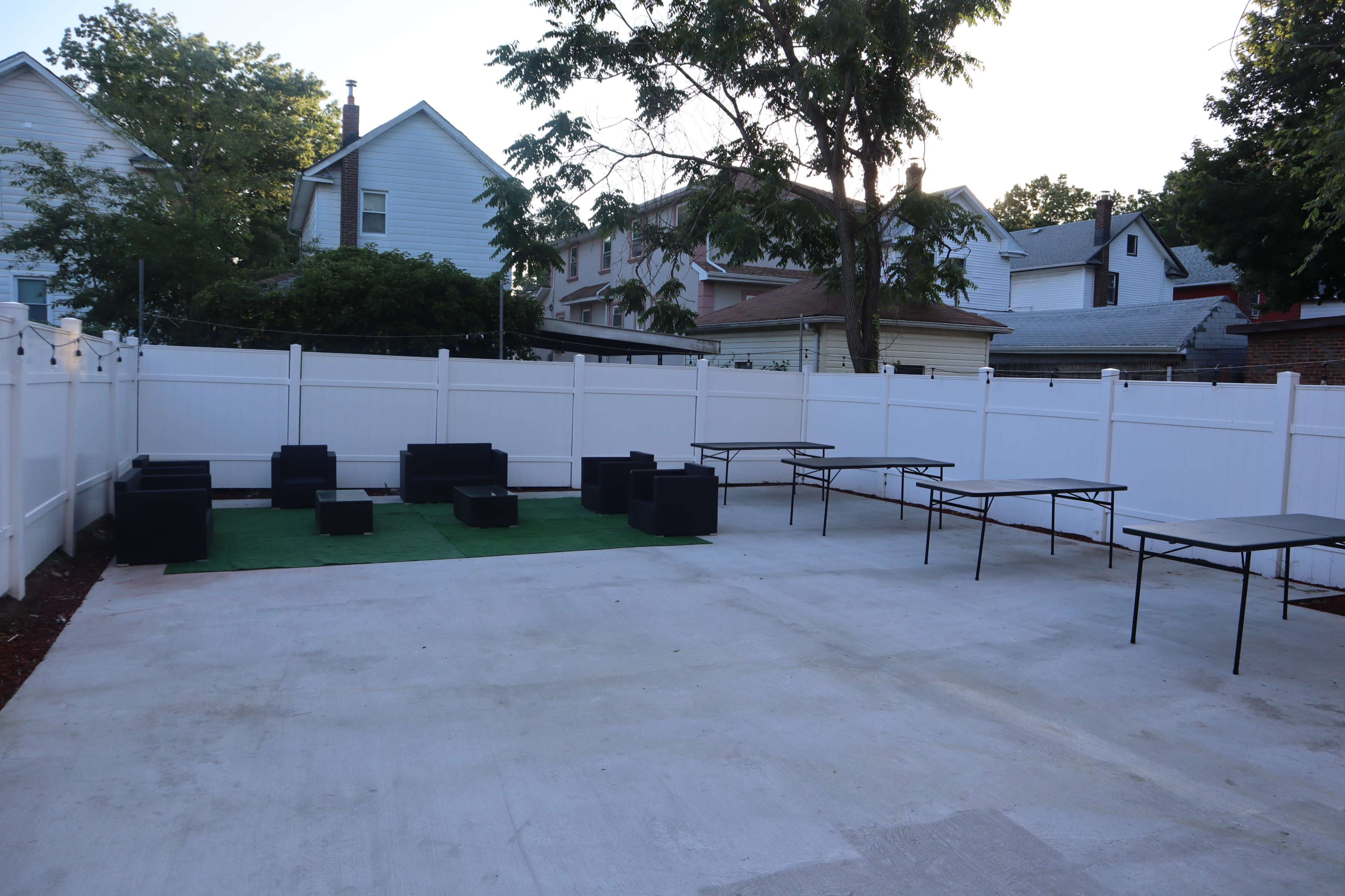 A spacious outdoor area enclosed by a white fence, featuring black seating cubes on green grass and several metal tables.