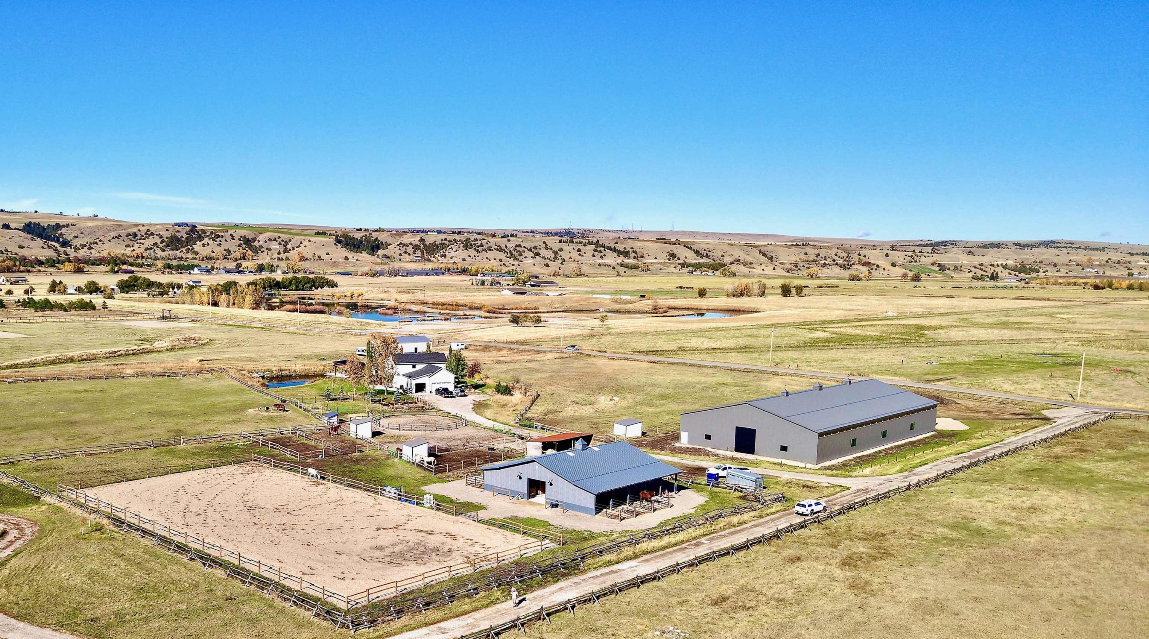 The image shows a spacious rural property with several buildings, including a barn, and fenced areas for horses, set against a backdrop of open fields and hills.