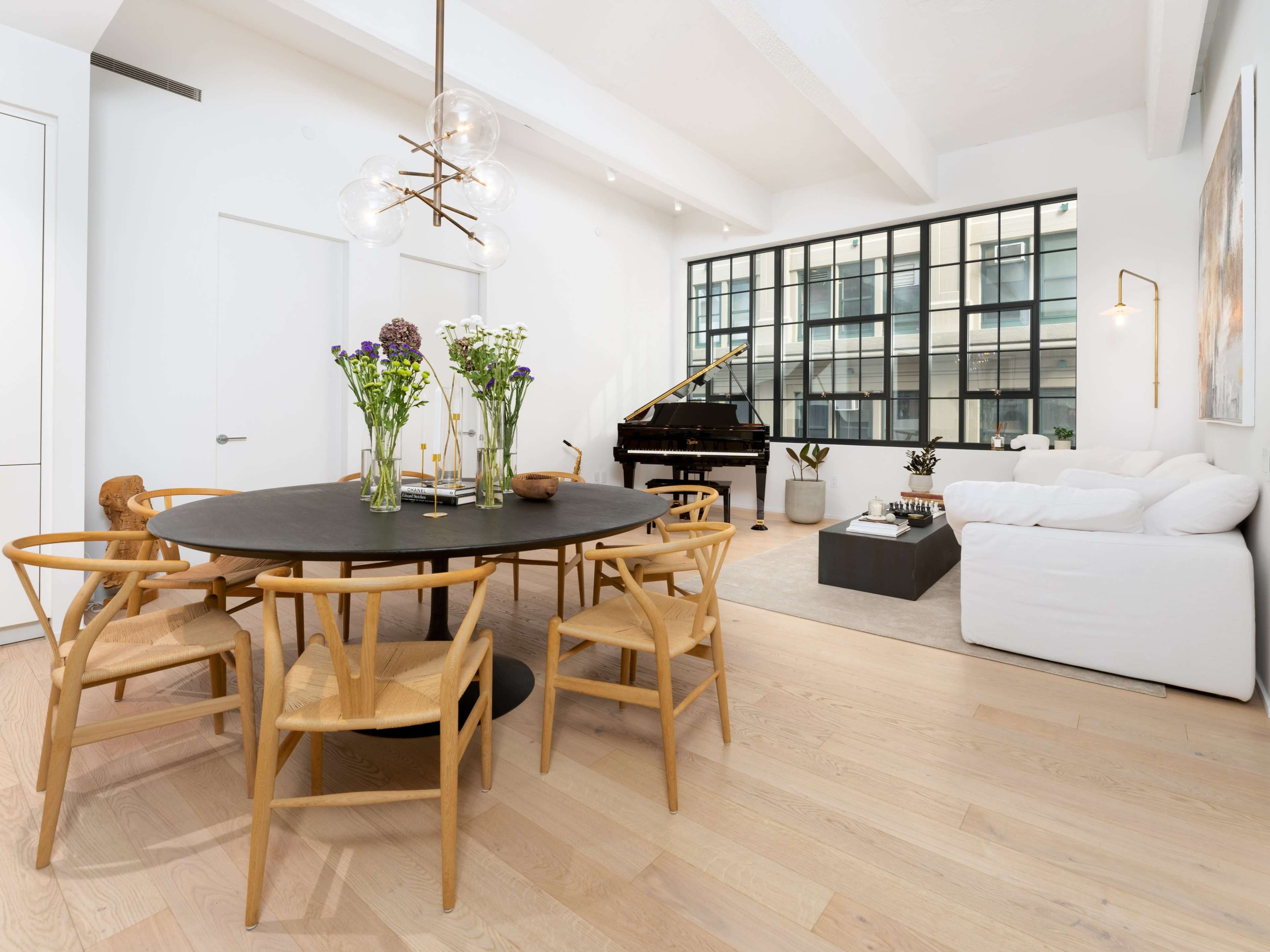 The image shows a modern living and dining space featuring a round black table with wooden chairs, a white sofa, a grand piano, and large windows allowing natural light.
