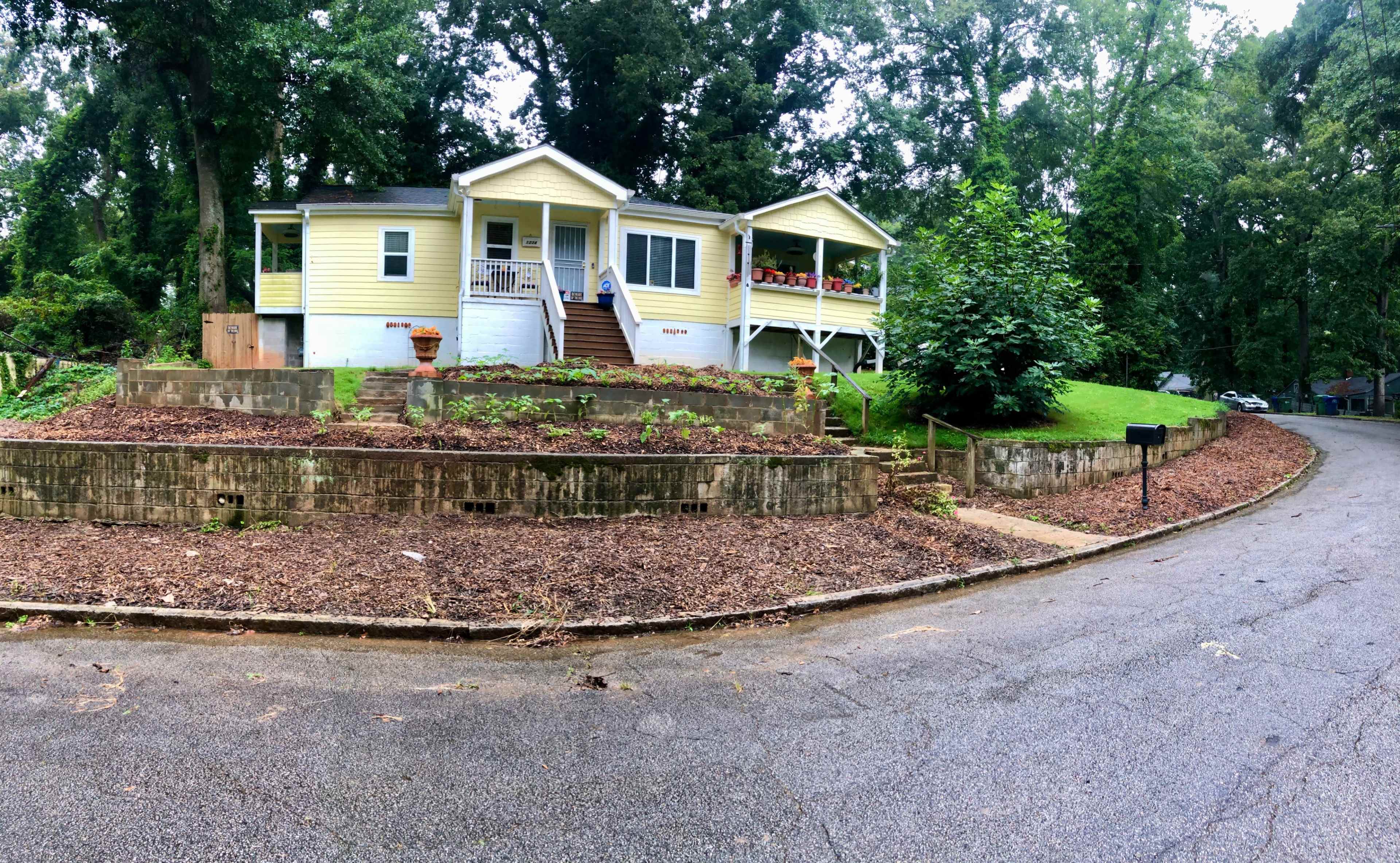 A yellow house with a porch and multiple windows sits on a landscaped lot at the end of a winding road, surrounded by trees.