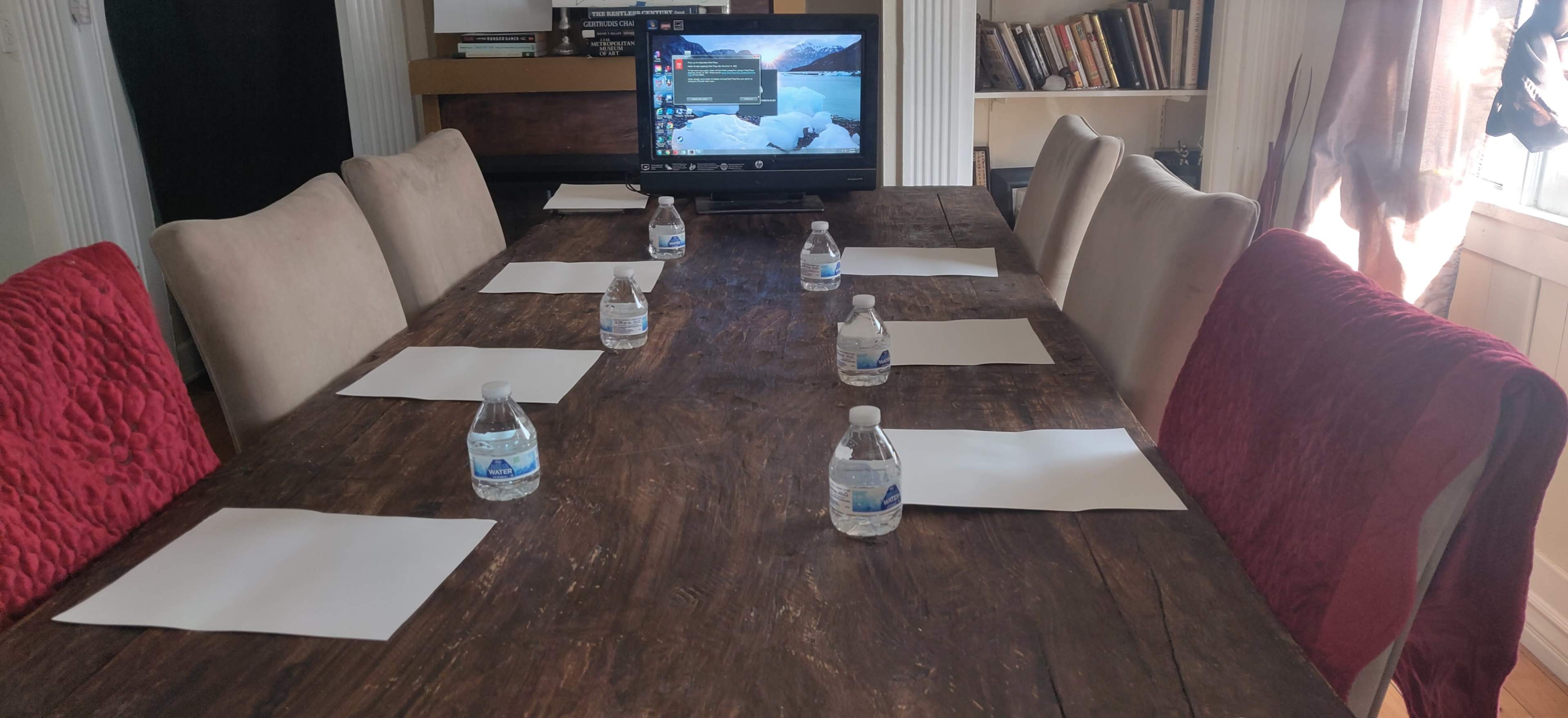A large wooden conference table is set with several bottles of water and blank sheets of paper, while a monitor displays a computer screen at one end.