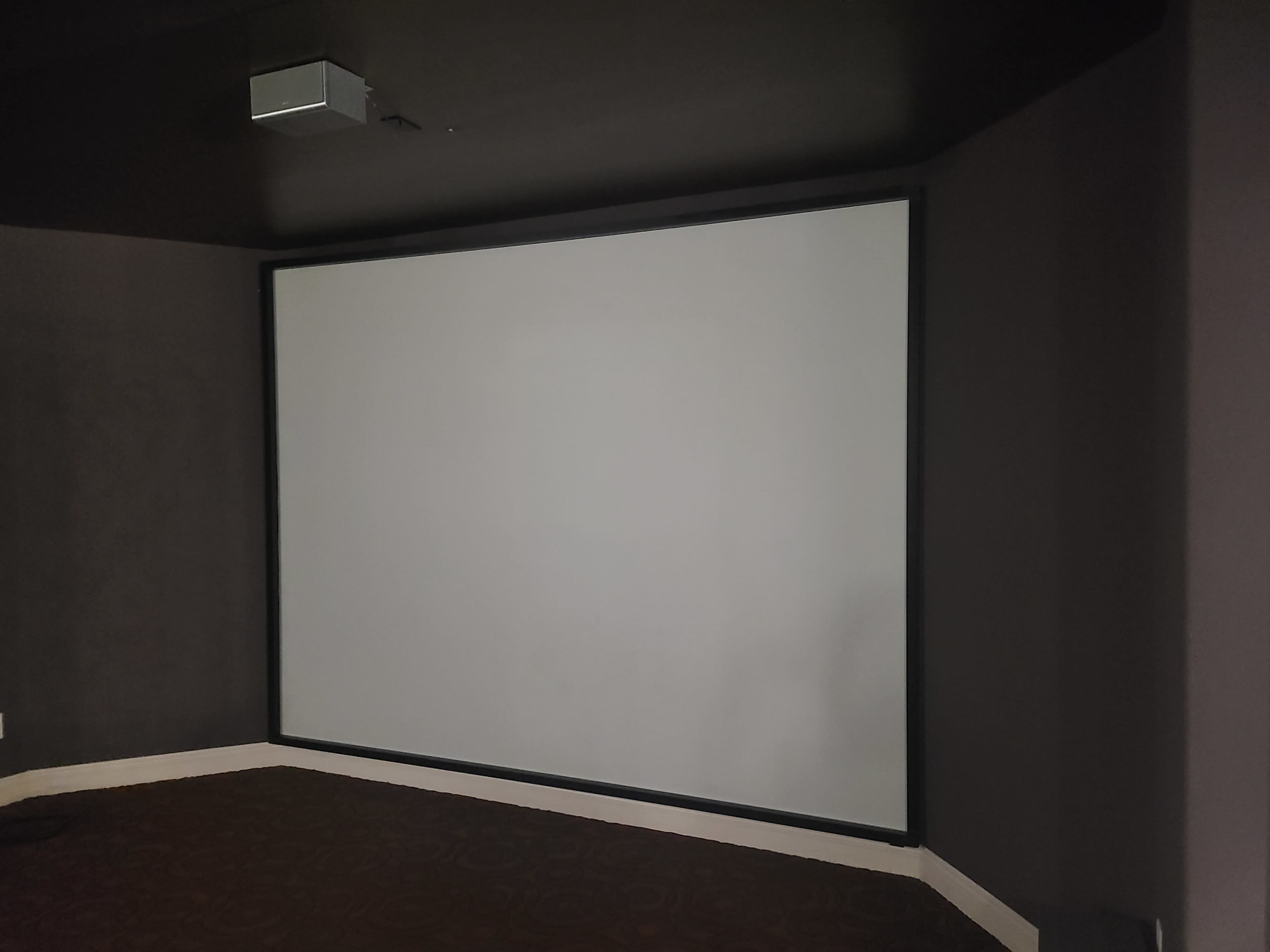 A blank projection screen mounted on a dark wall in a dimly lit room.