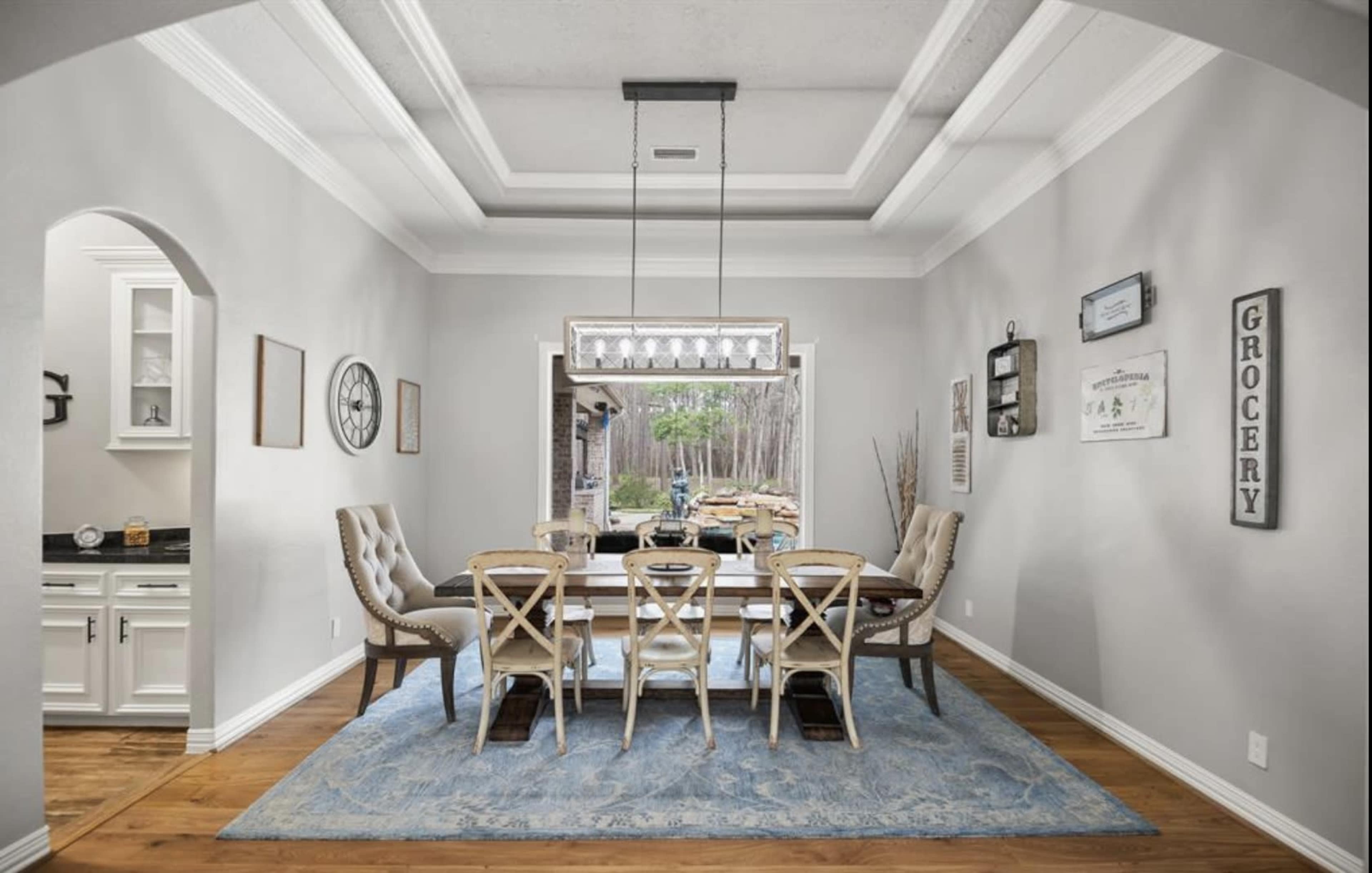 A dining room features a large wooden table surrounded by six chairs, with a chandelier overhead and light gray walls.