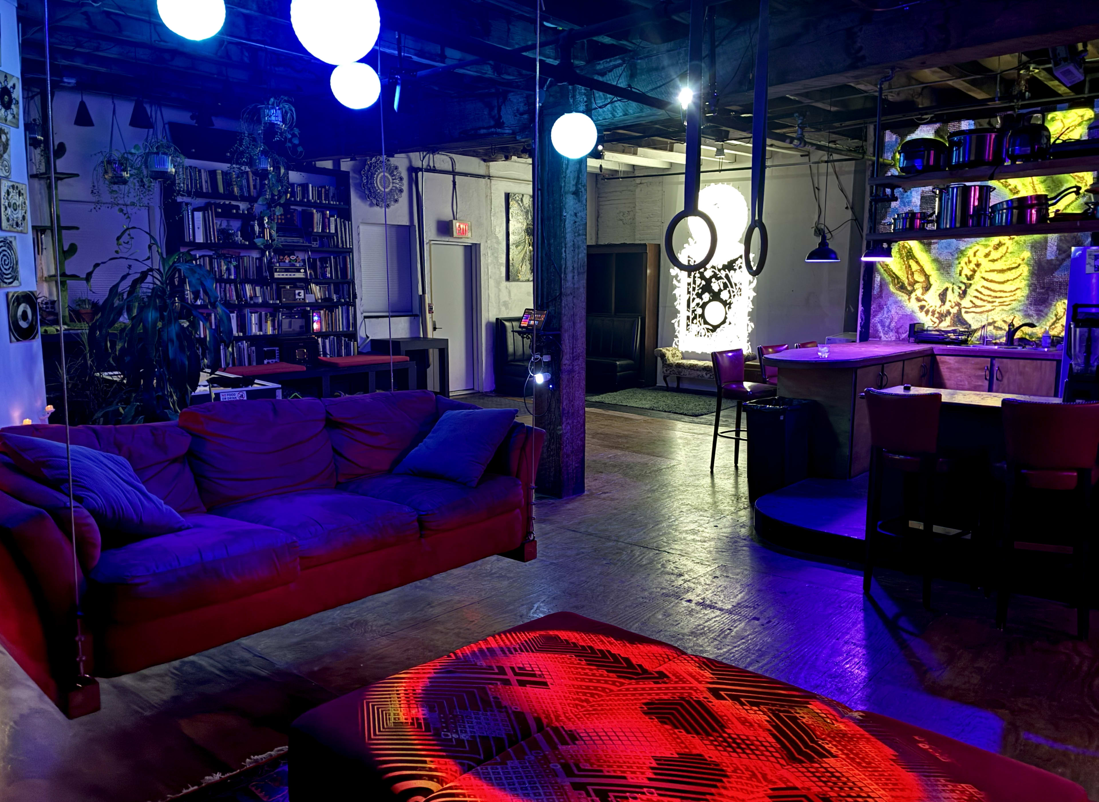 The image shows a dimly lit indoor space featuring a red sofa, decorative lighting, a bookshelf, and an illuminated bar area in the background.