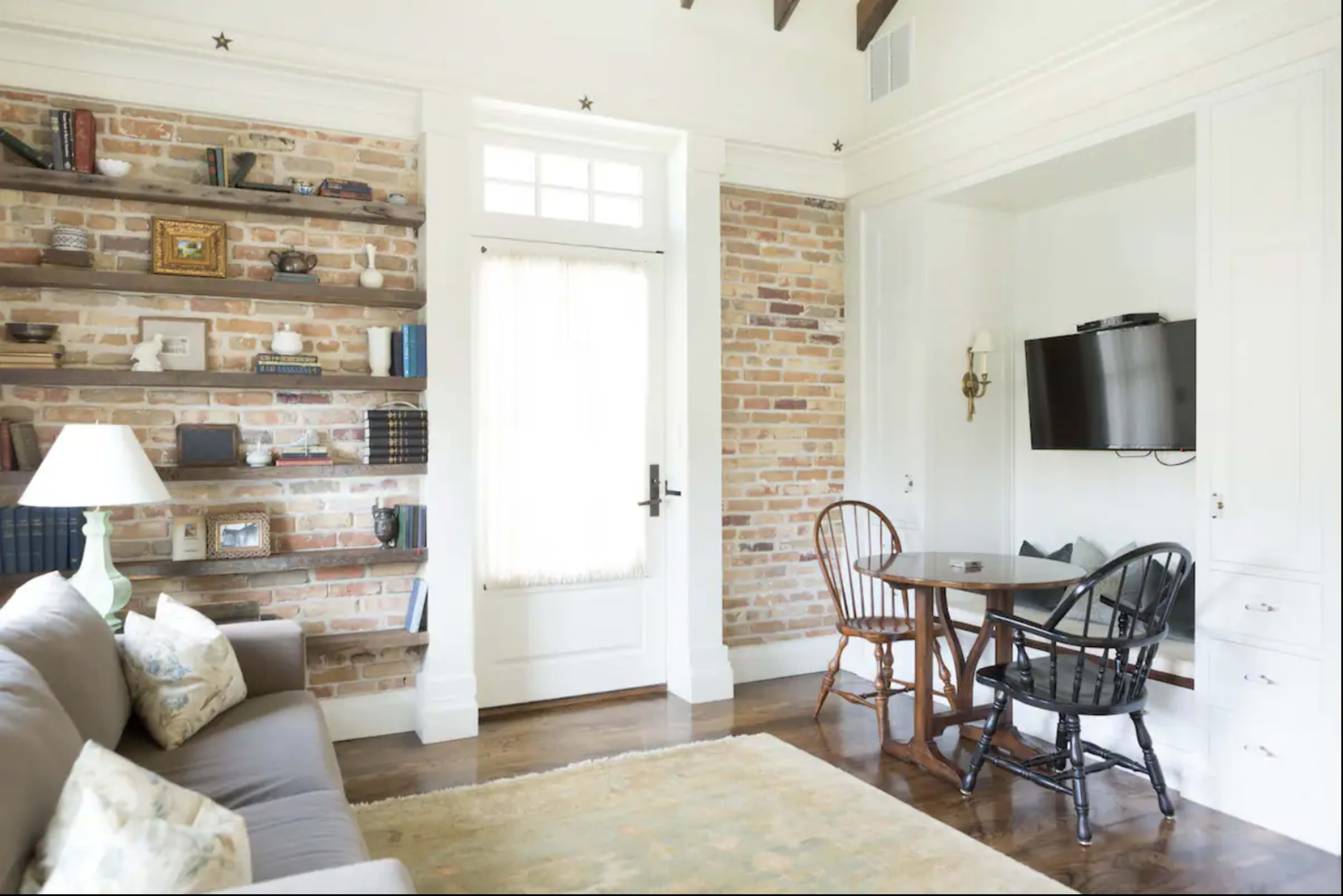A living room features a gray sofa, a circular dining table with two chairs, exposed brick walls, and a mounted television.
