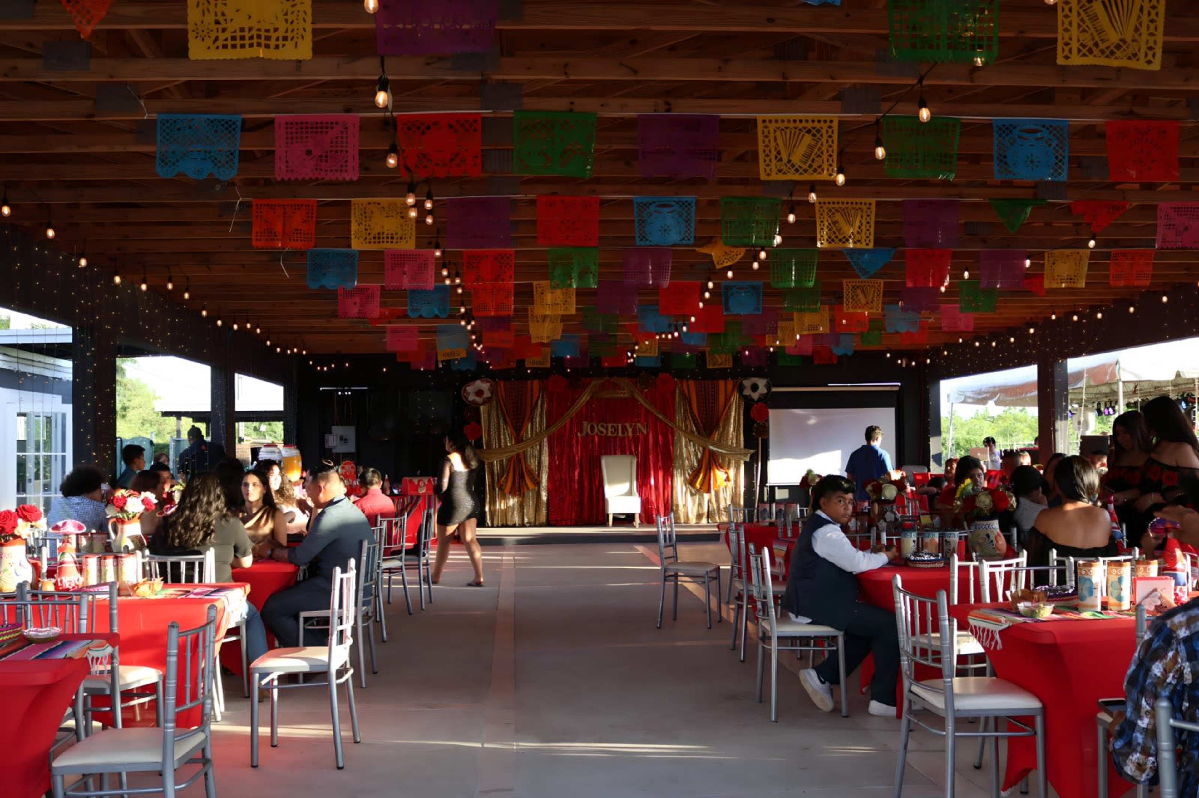 A festive event space is decorated with colorful papel picado banners, featuring red table settings and soft lighting, ready for a celebration.