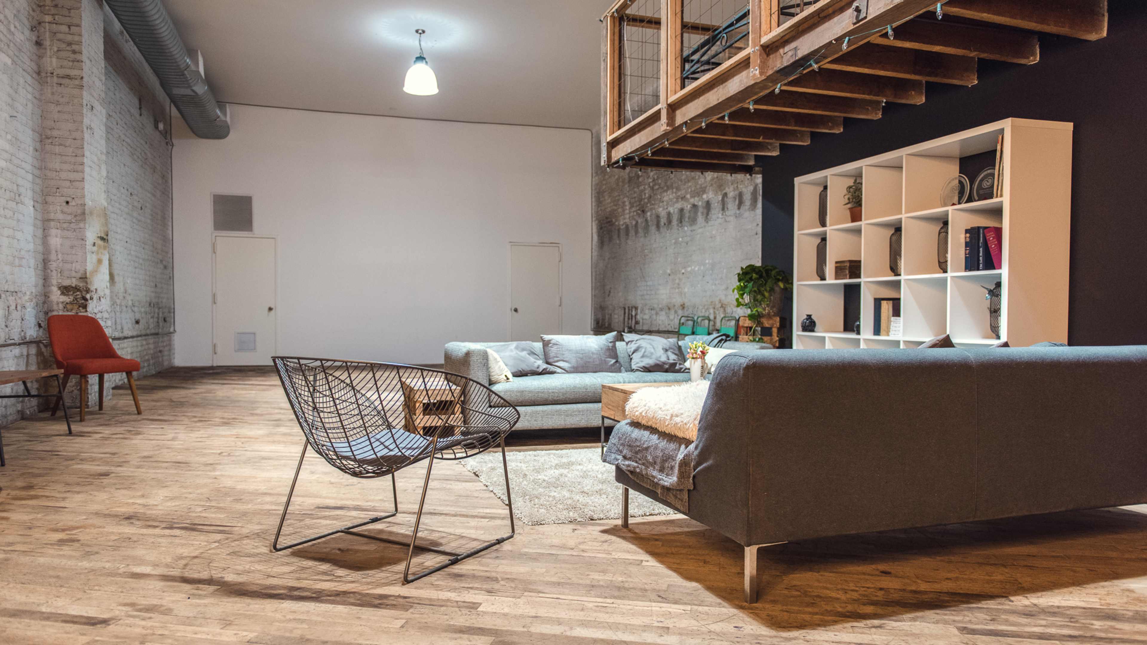 A spacious, minimalist room with wooden flooring, featuring a seating area with a sofa, a lounge chair, a coffee table, and a bookshelf against a black wall.