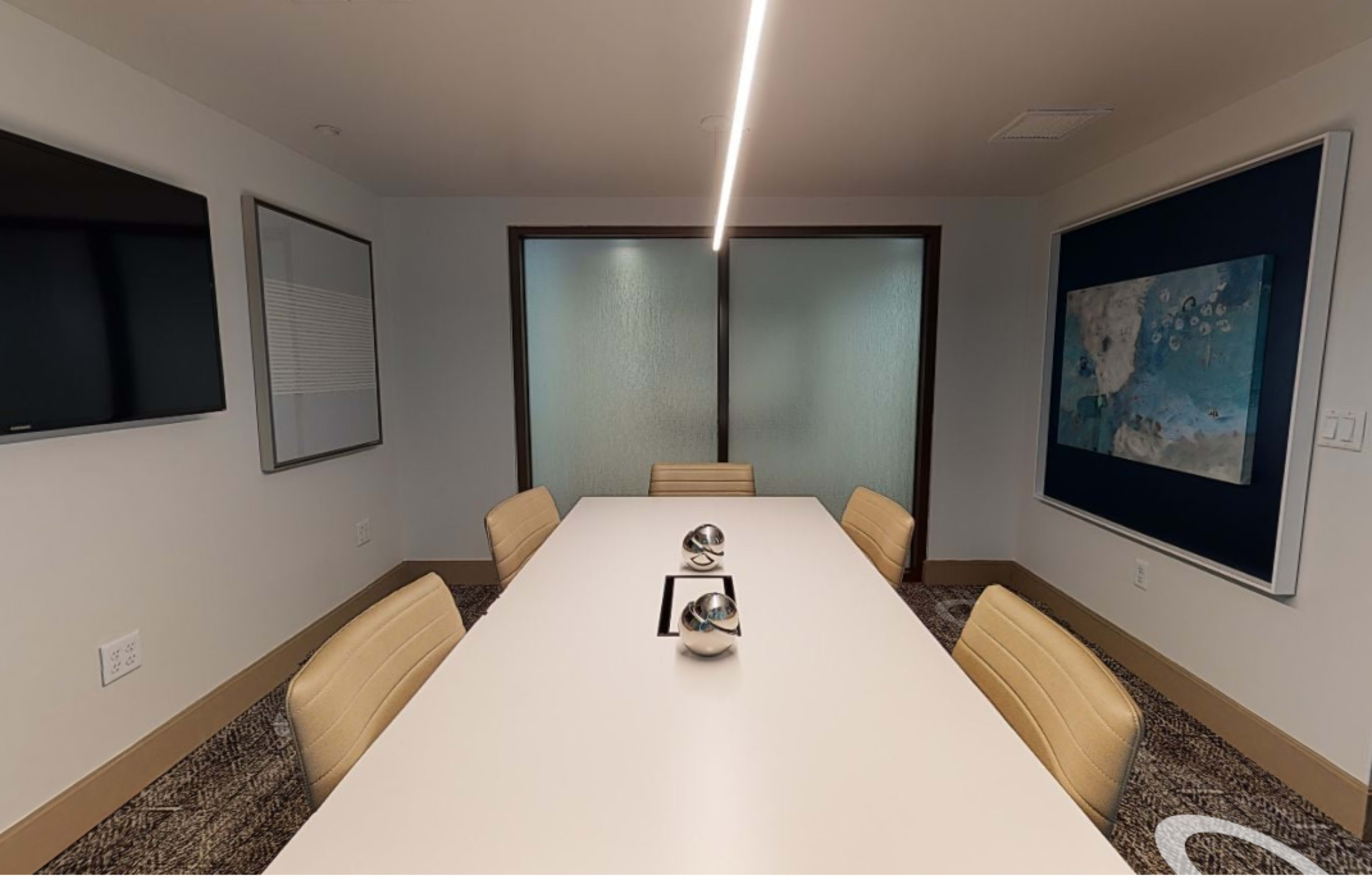 A modern conference room with a rectangular table, four beige chairs, a wall-mounted TV, and frosted glass doors.