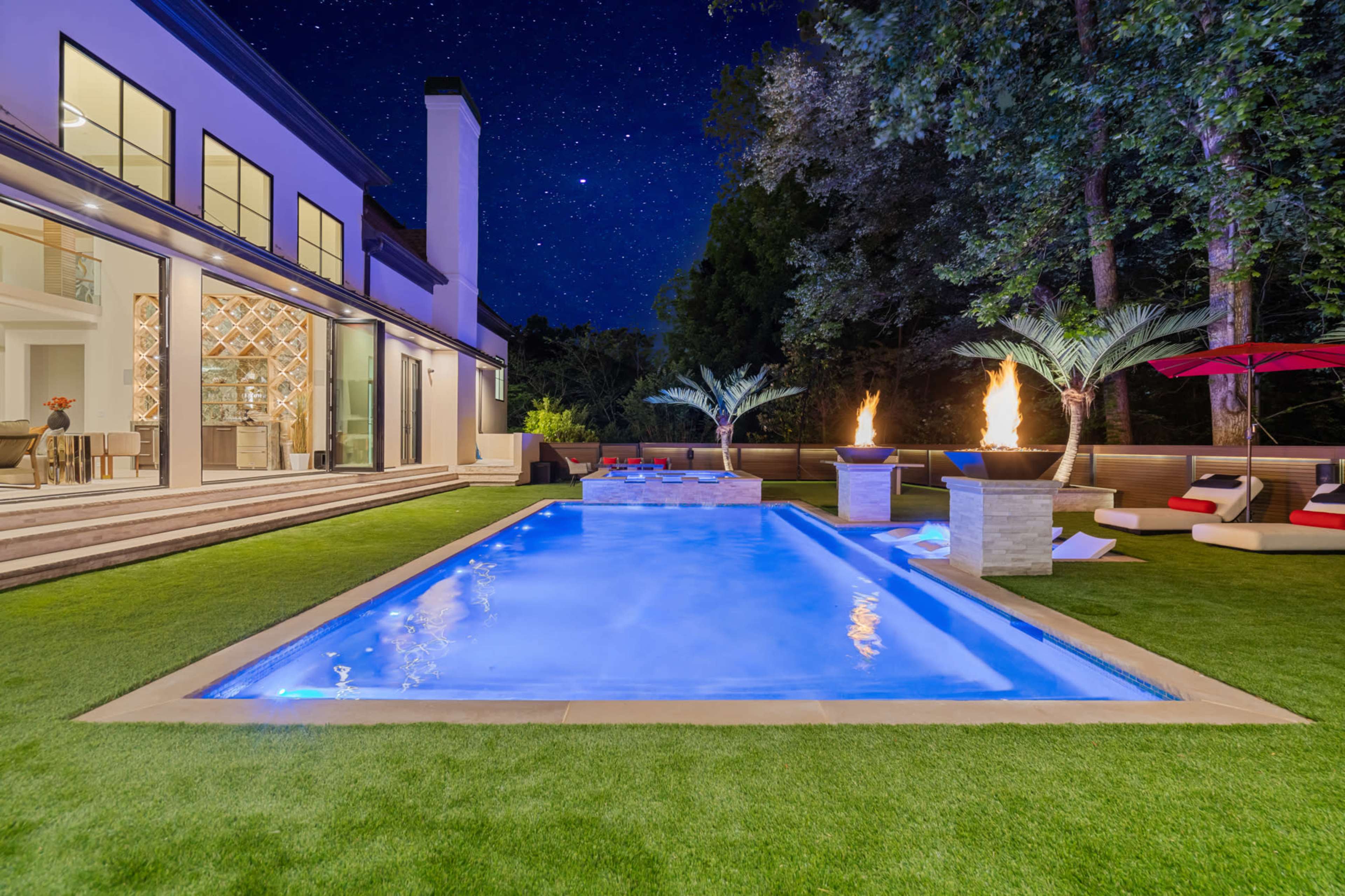 A well-lit modern pool area features a rectangular swimming pool surrounded by green grass, lounge chairs, and palm trees under a starry night sky.