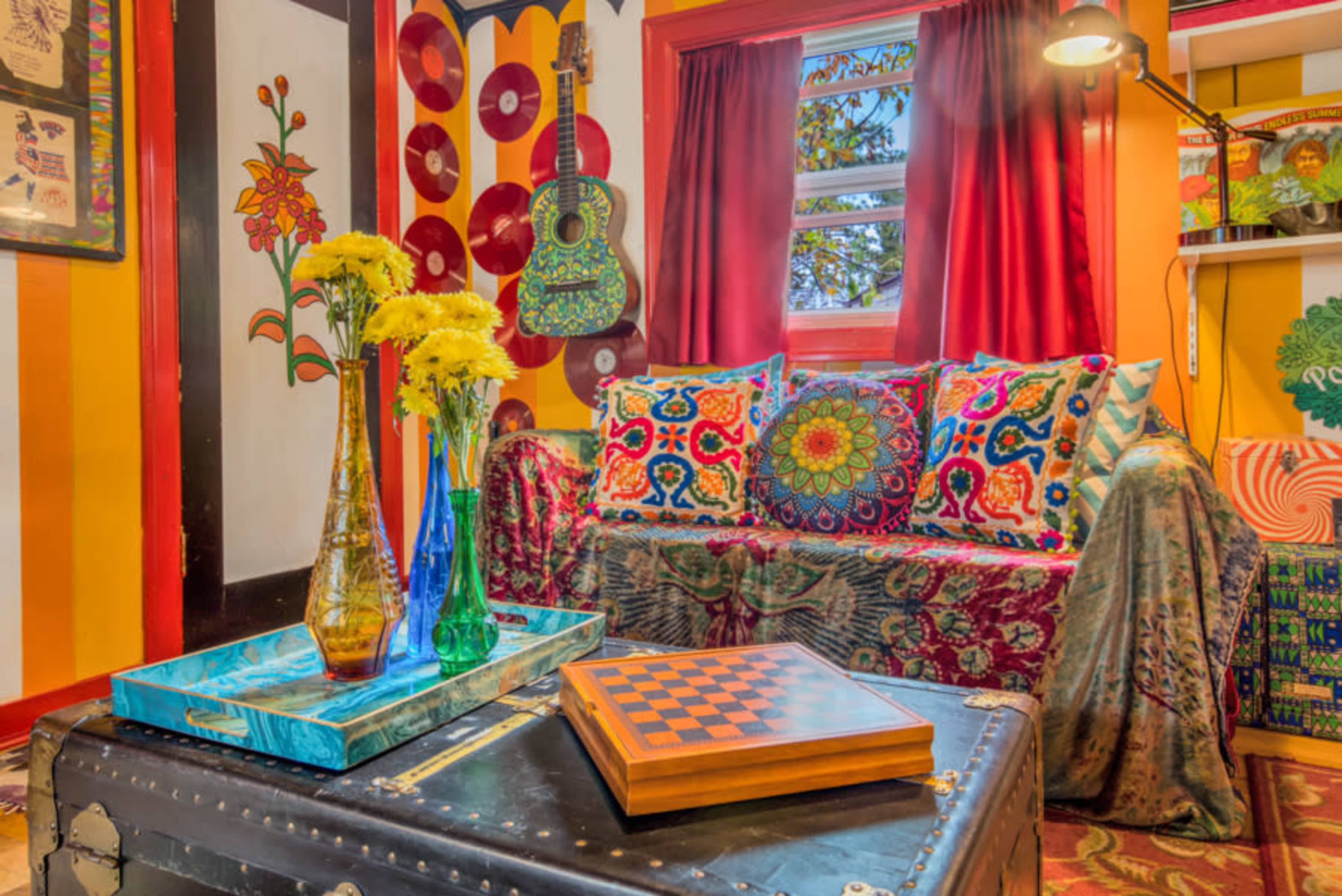 The image features a vibrantly decorated room with a patterned couch, colorful pillows, a coffee table with a checkered game board, and decorative flowers in glass vases.