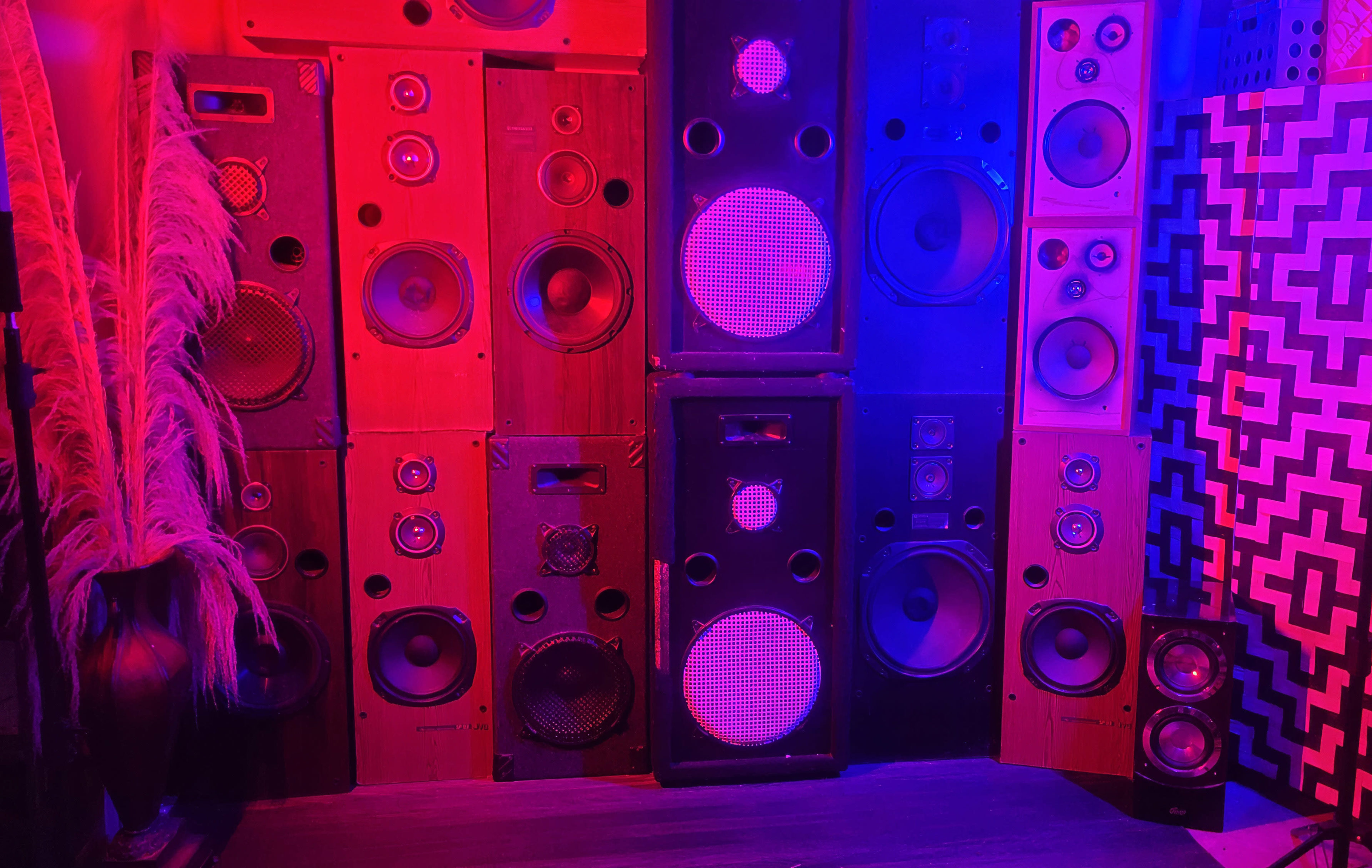 The image shows a wall adorned with a collection of various loudspeakers and amplifiers, lit in vibrant red and blue hues.