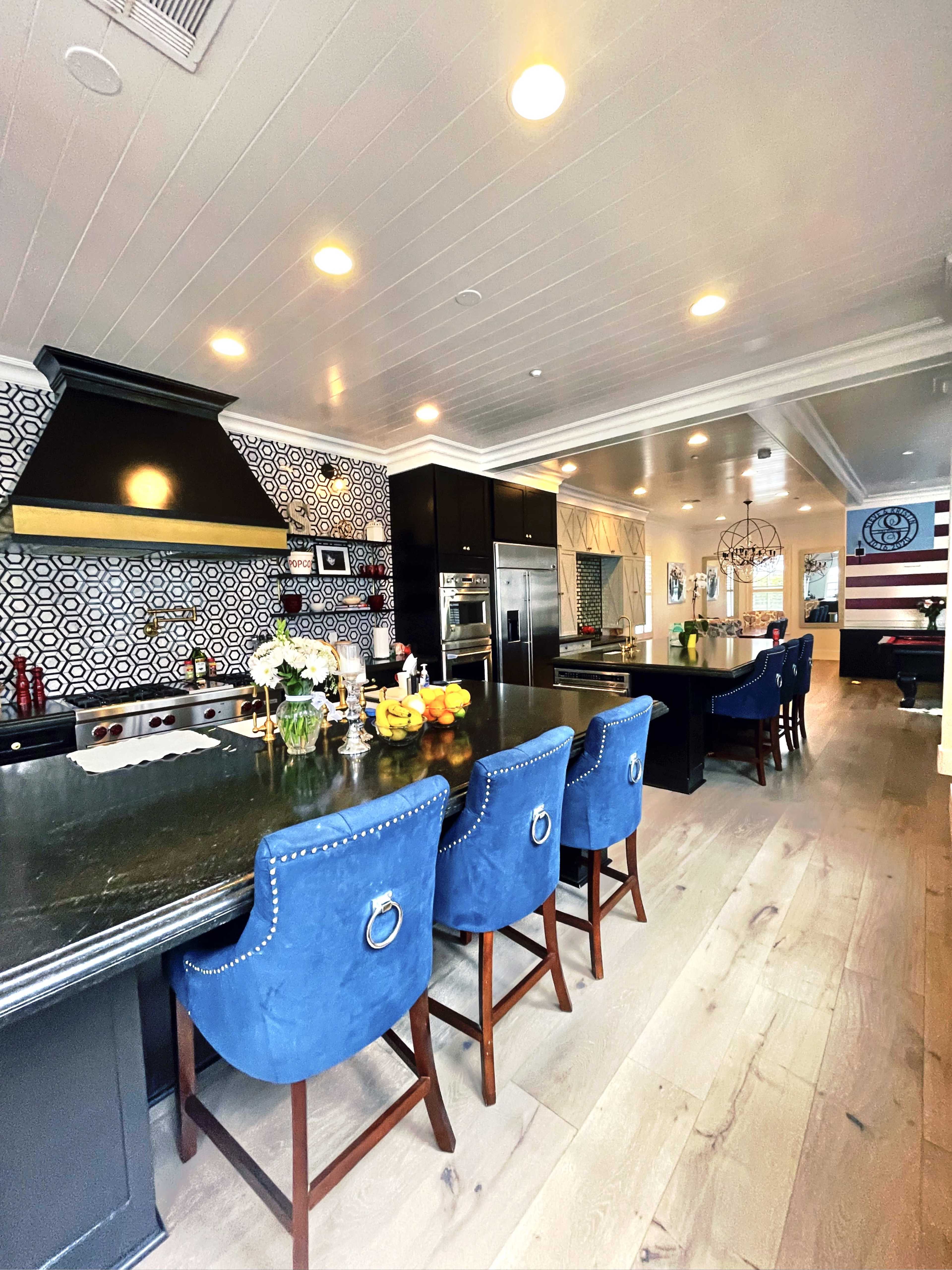 A modern kitchen features a black stone island with blue upholstered chairs, patterned wallpaper, and stainless steel appliances.
