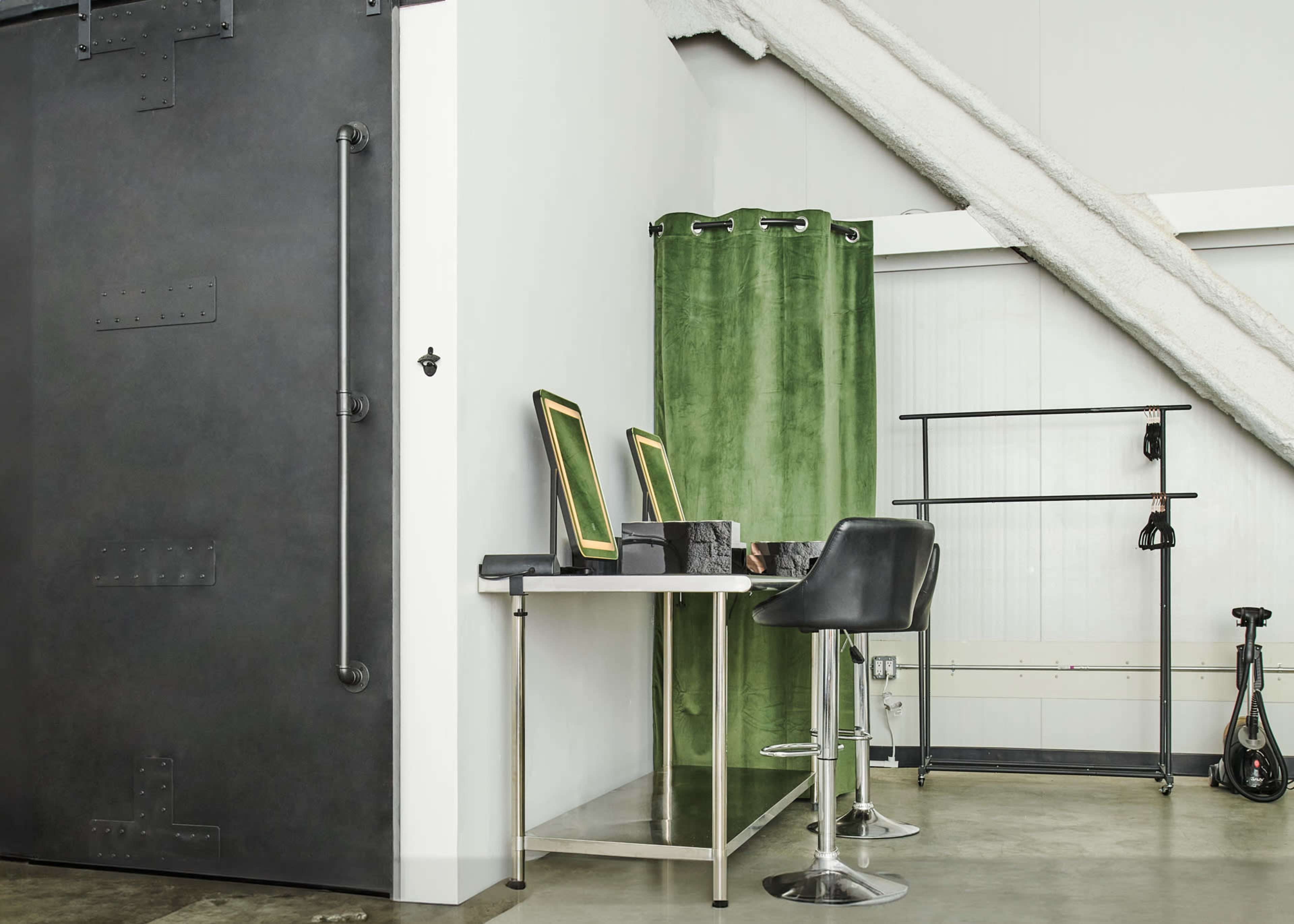 The image shows a modern interior space featuring a black metal door, a green curtain, a metal table with mirrors, and a stool, along with a clothing rack and a bicycle in the background.