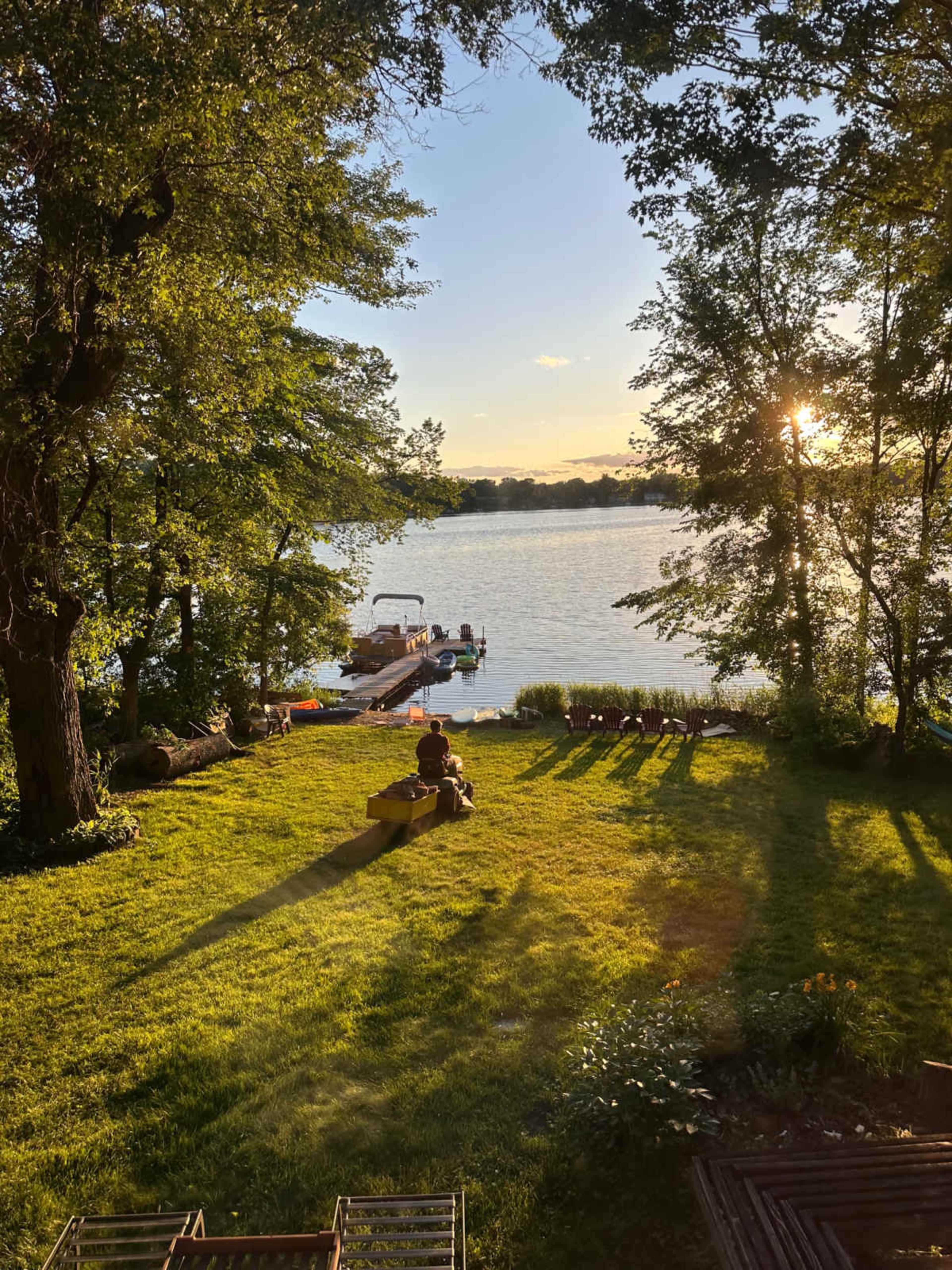 A person sits on the grass near a lake at sunset, with a dock and boats visible in the background.