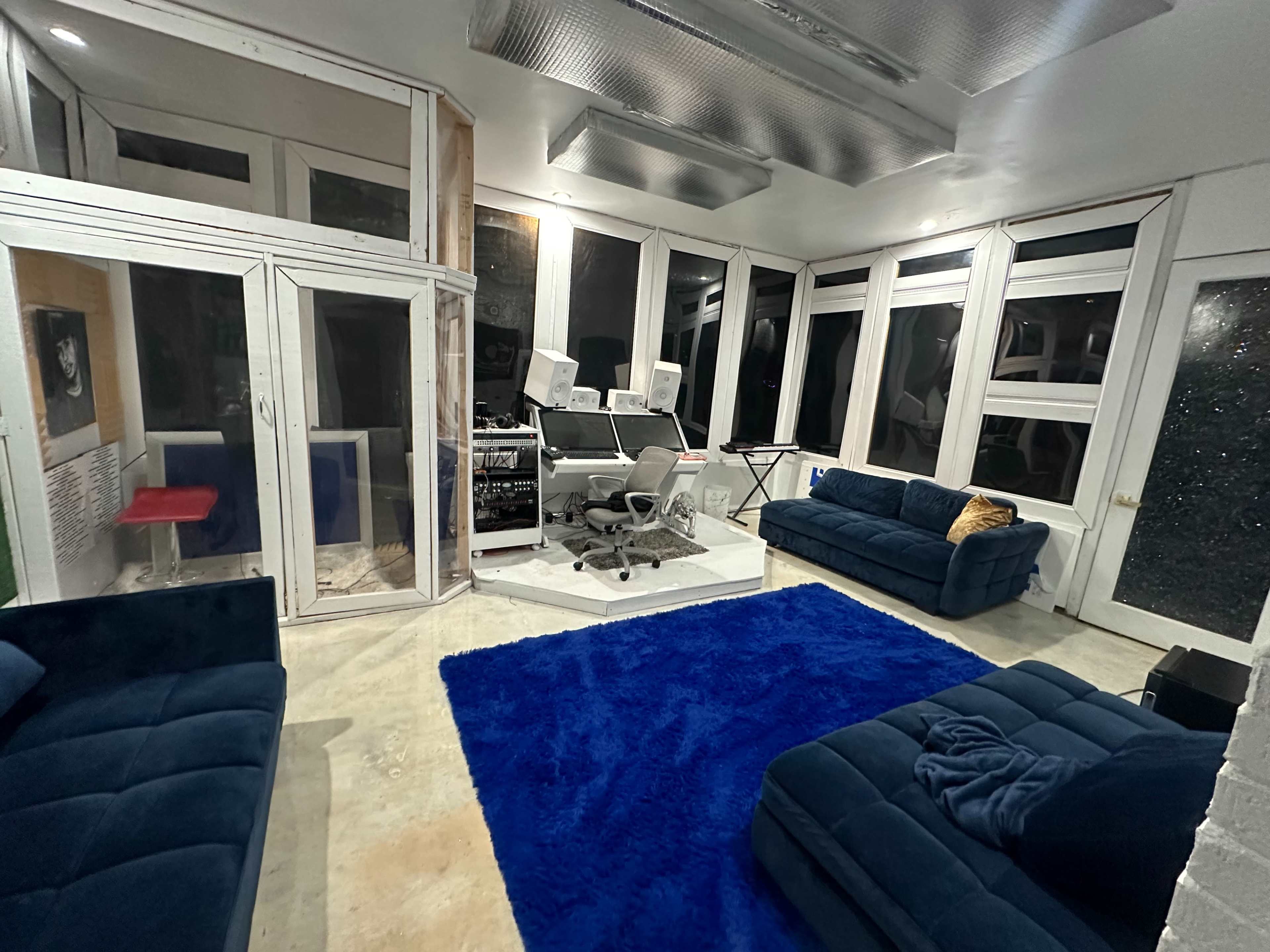 The room features large windows with a view, a blue carpet, and two blue couches arranged around a central area with modern electronic equipment.