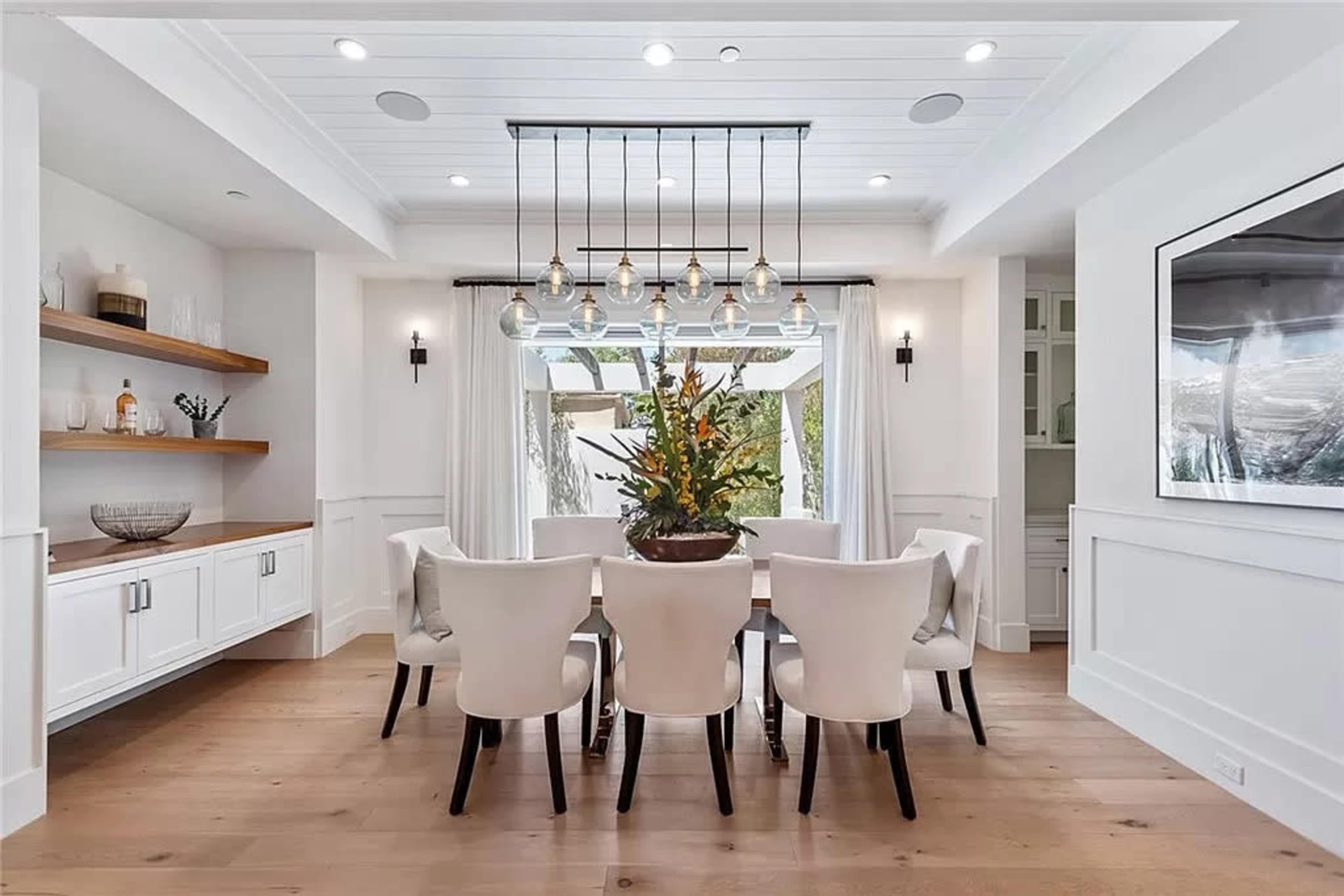 A spacious dining room features a long table surrounded by eight upholstered chairs, with a decorative floral arrangement and pendant lighting above.