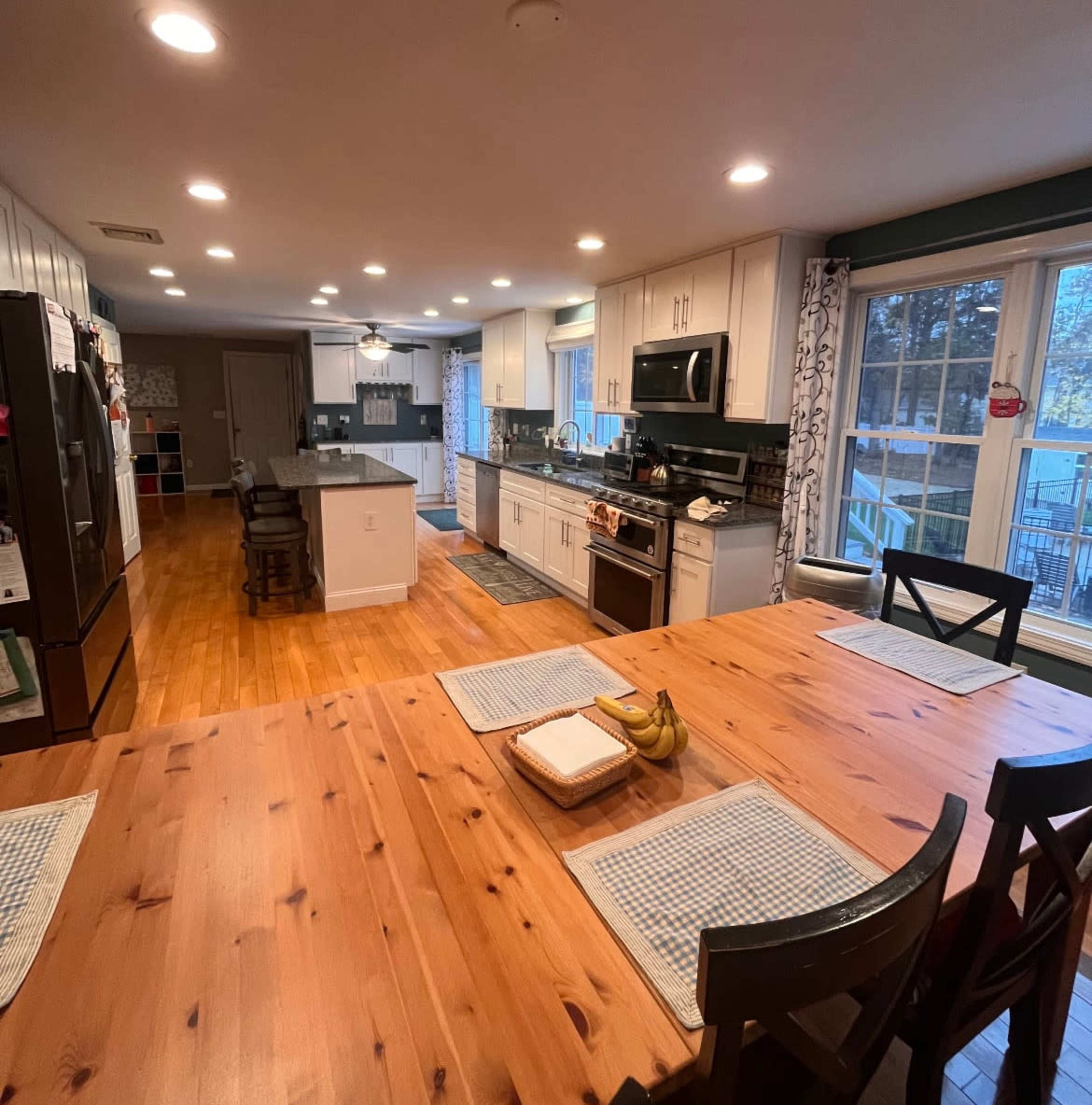 The image shows a modern kitchen and dining area with a wooden table, black chairs, and a well-equipped kitchen featuring stainless steel appliances and ample cabinetry.