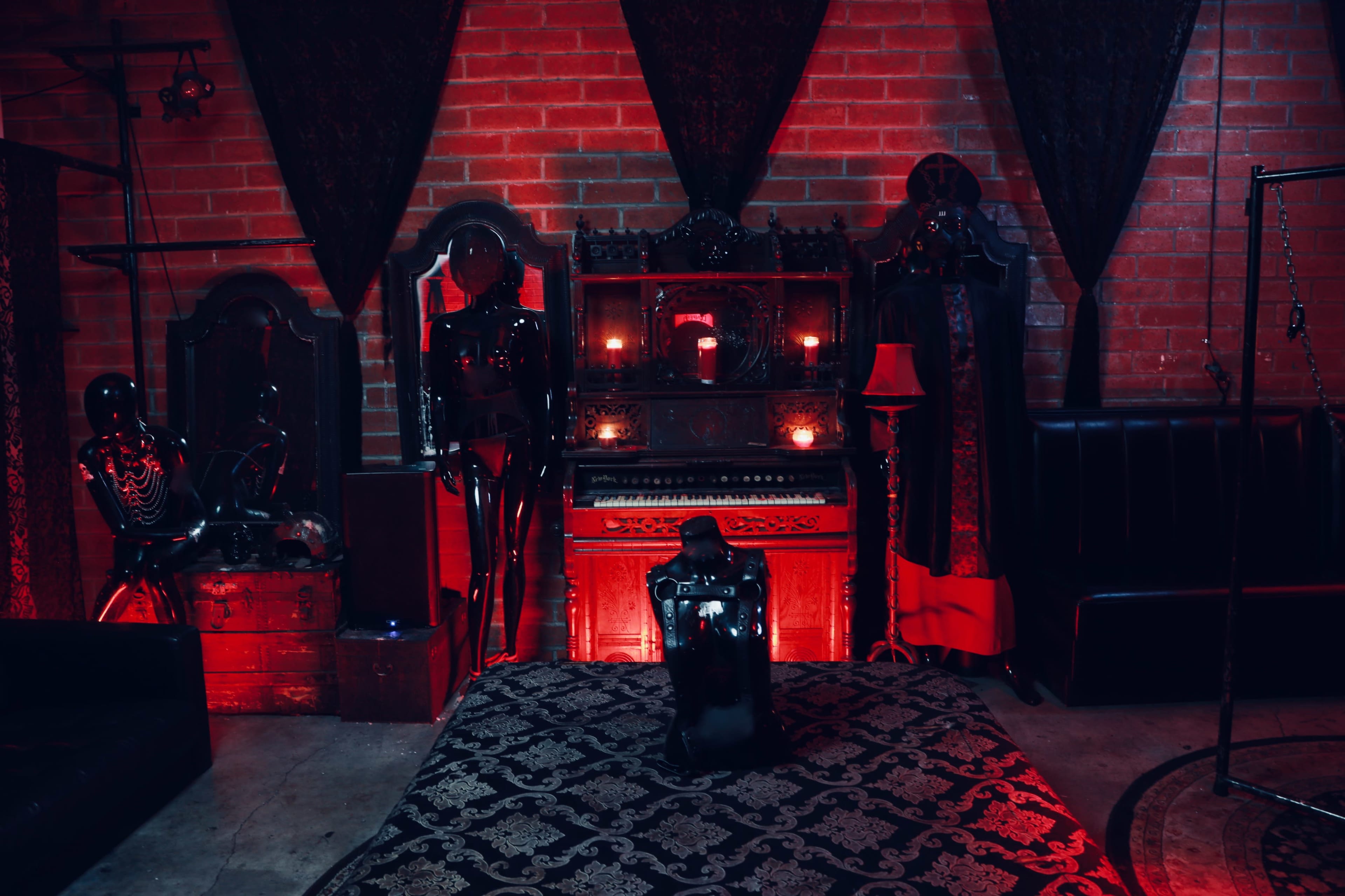 The scene features a dimly lit room with red lighting, showcasing an ornate piano surrounded by vintage furniture and mannequins adorned in dark attire.