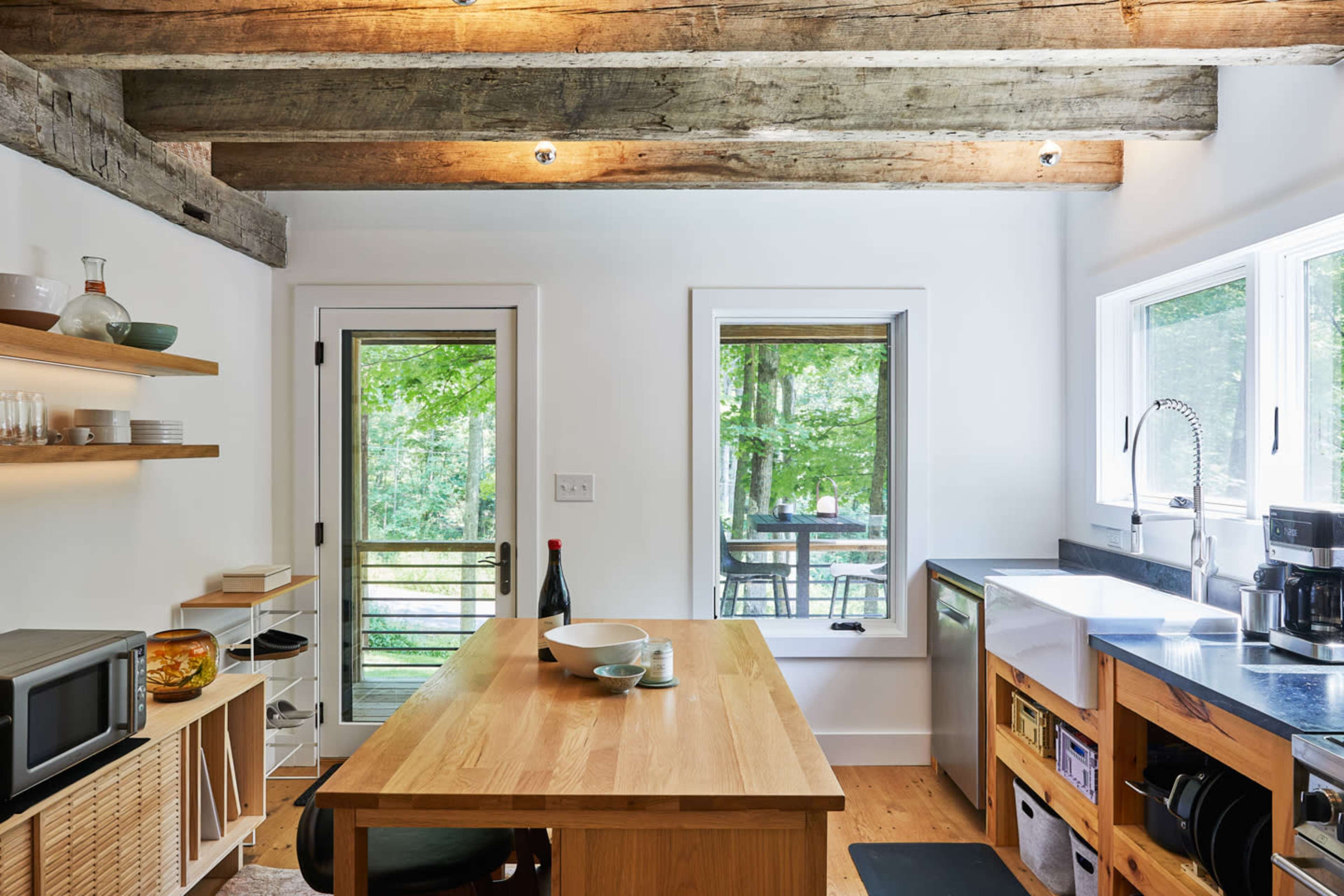 A modern kitchen features wooden beams, an island table, open shelving, and views of a forested outdoor deck through large windows.