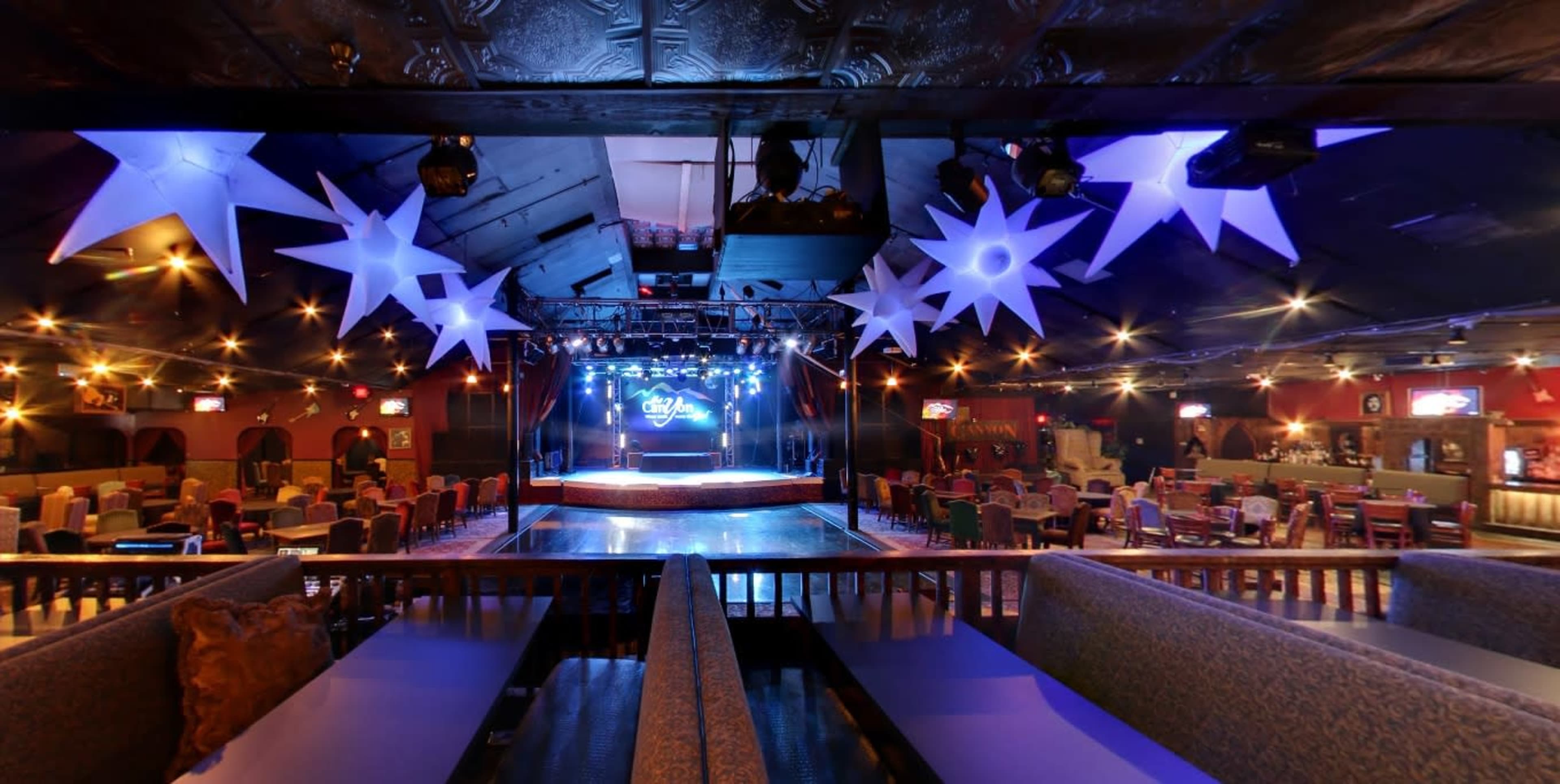 A spacious entertainment venue with a stage, illuminated by colorful lights, surrounded by seating areas and decorations.