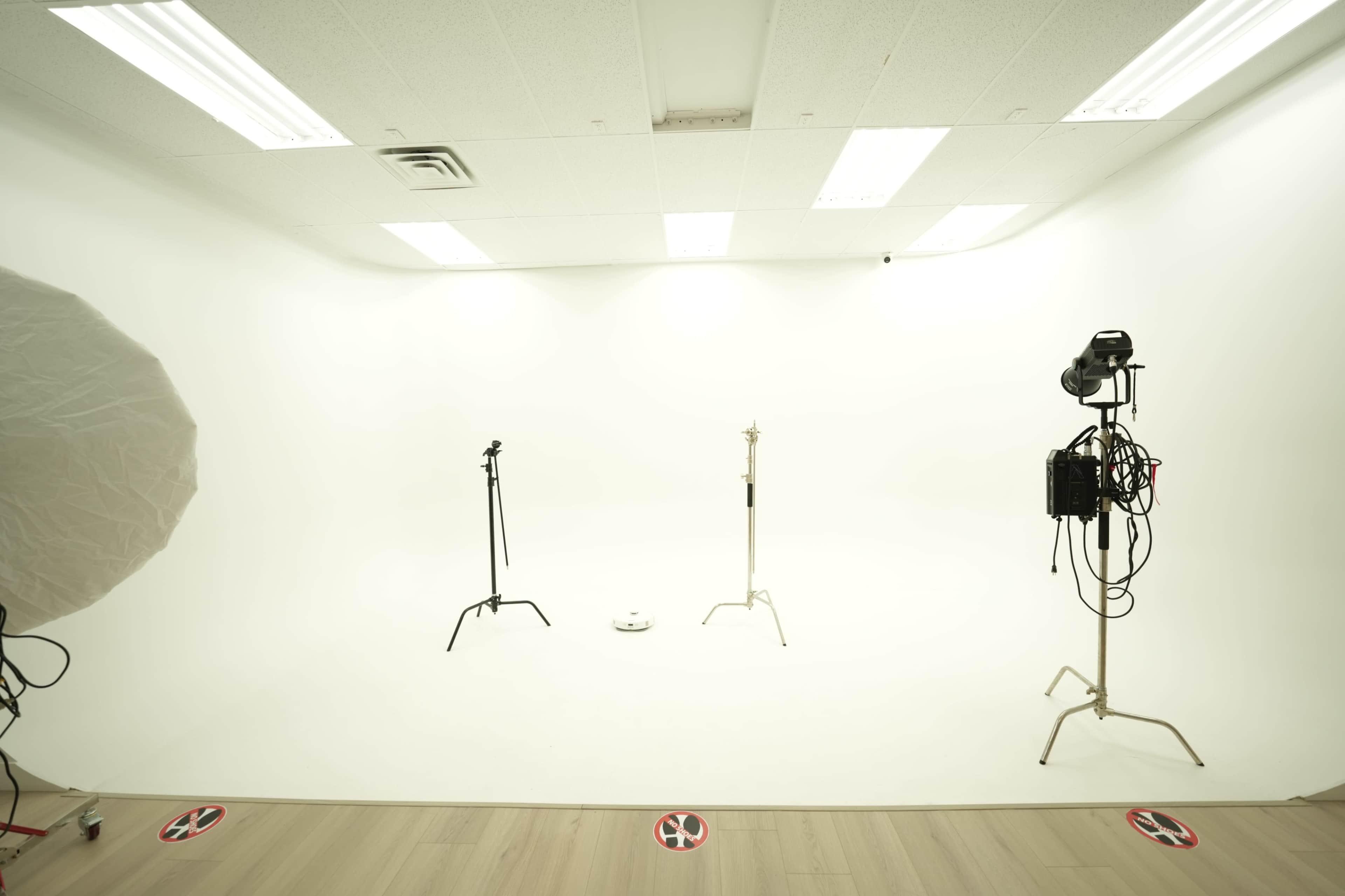 The image shows a minimalistic photography studio with three stands and studio lighting equipment set against a white backdrop.