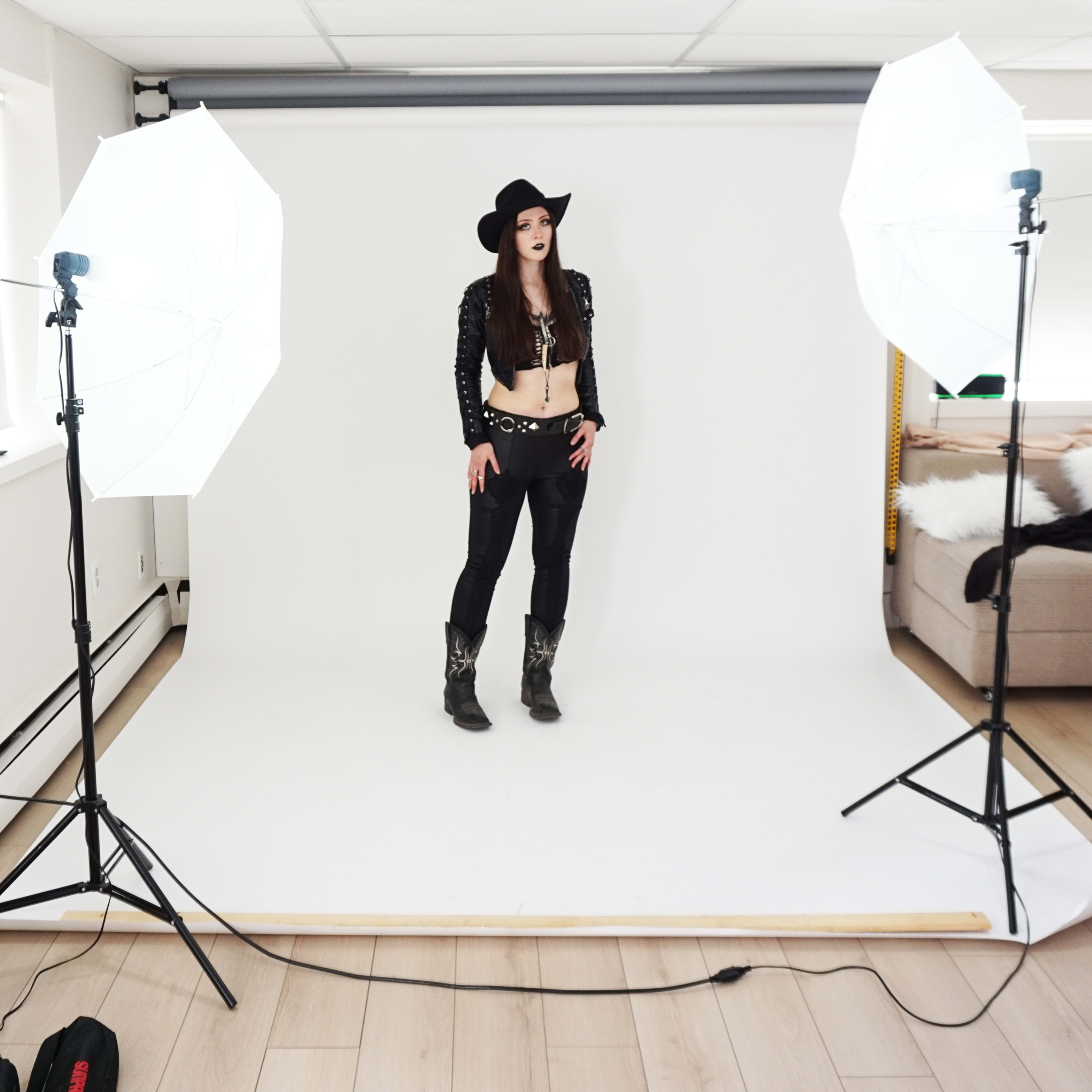 A model poses on a white backdrop in a photography studio, flanked by two large lighting umbrellas.