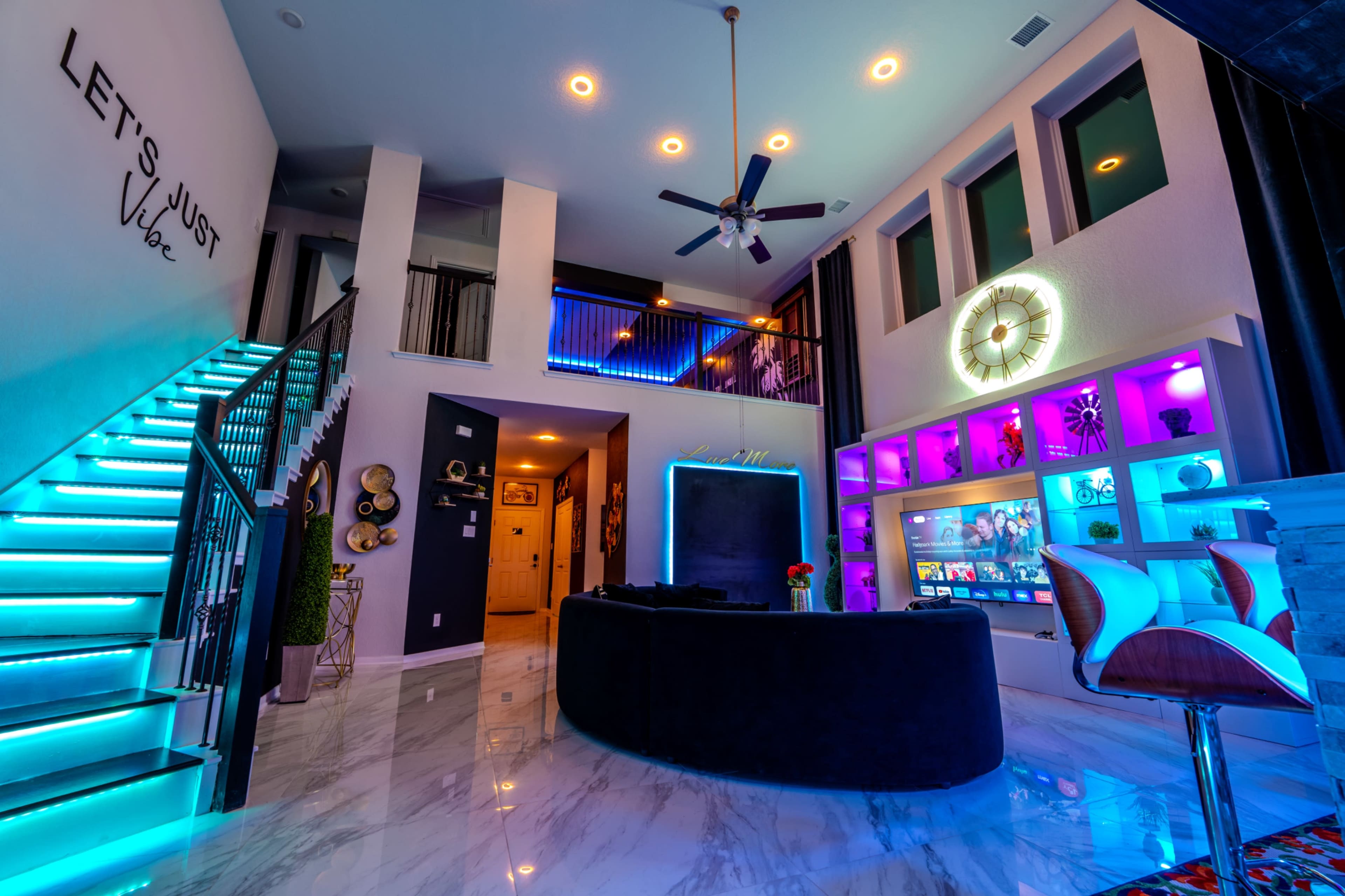 The image shows a modern living space featuring a sleek black couch, colorful LED lighting, and a staircase leading to an upper level.
