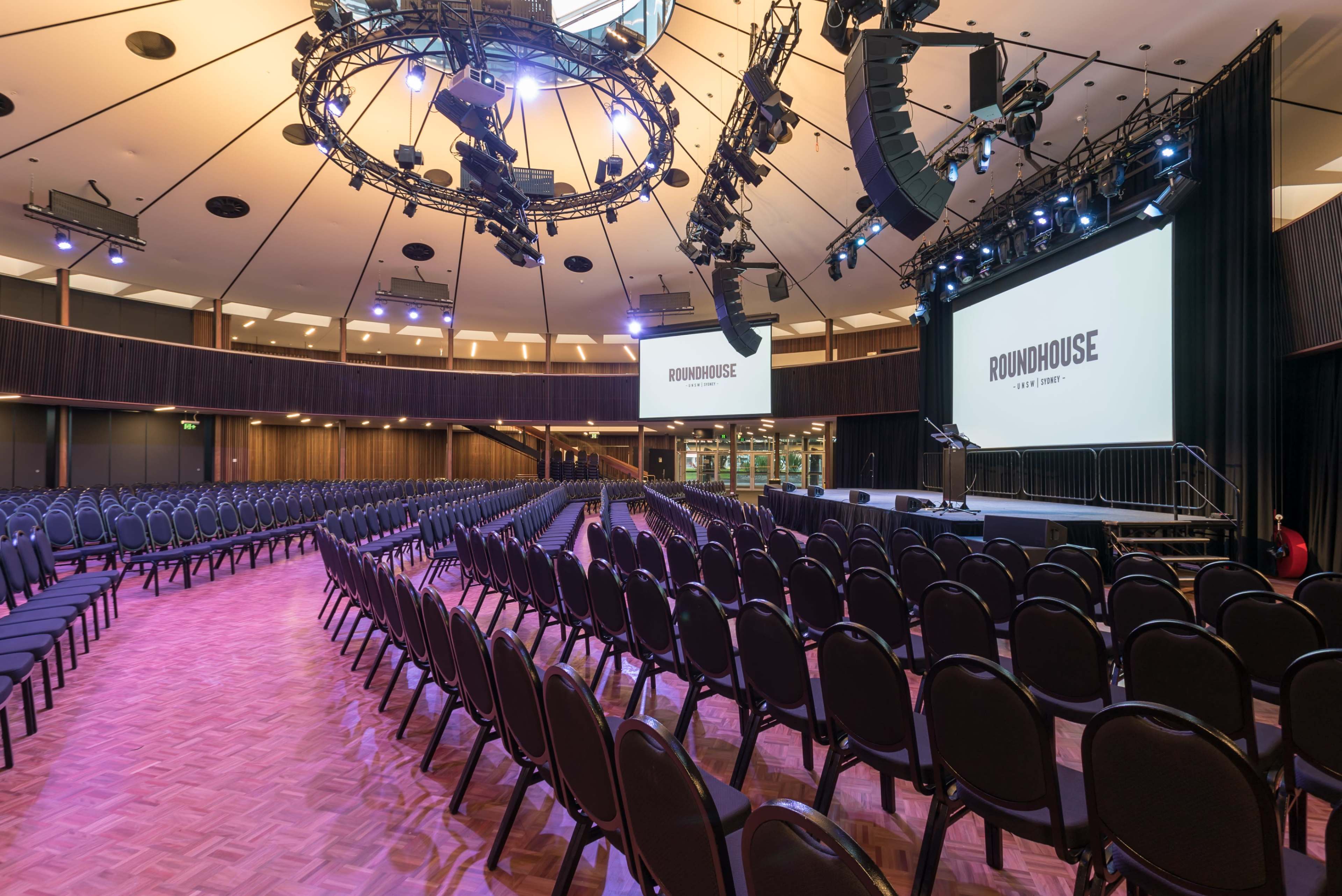 A large indoor auditorium with rows of black chairs facing a stage, illuminated by overhead lighting in a circular room.