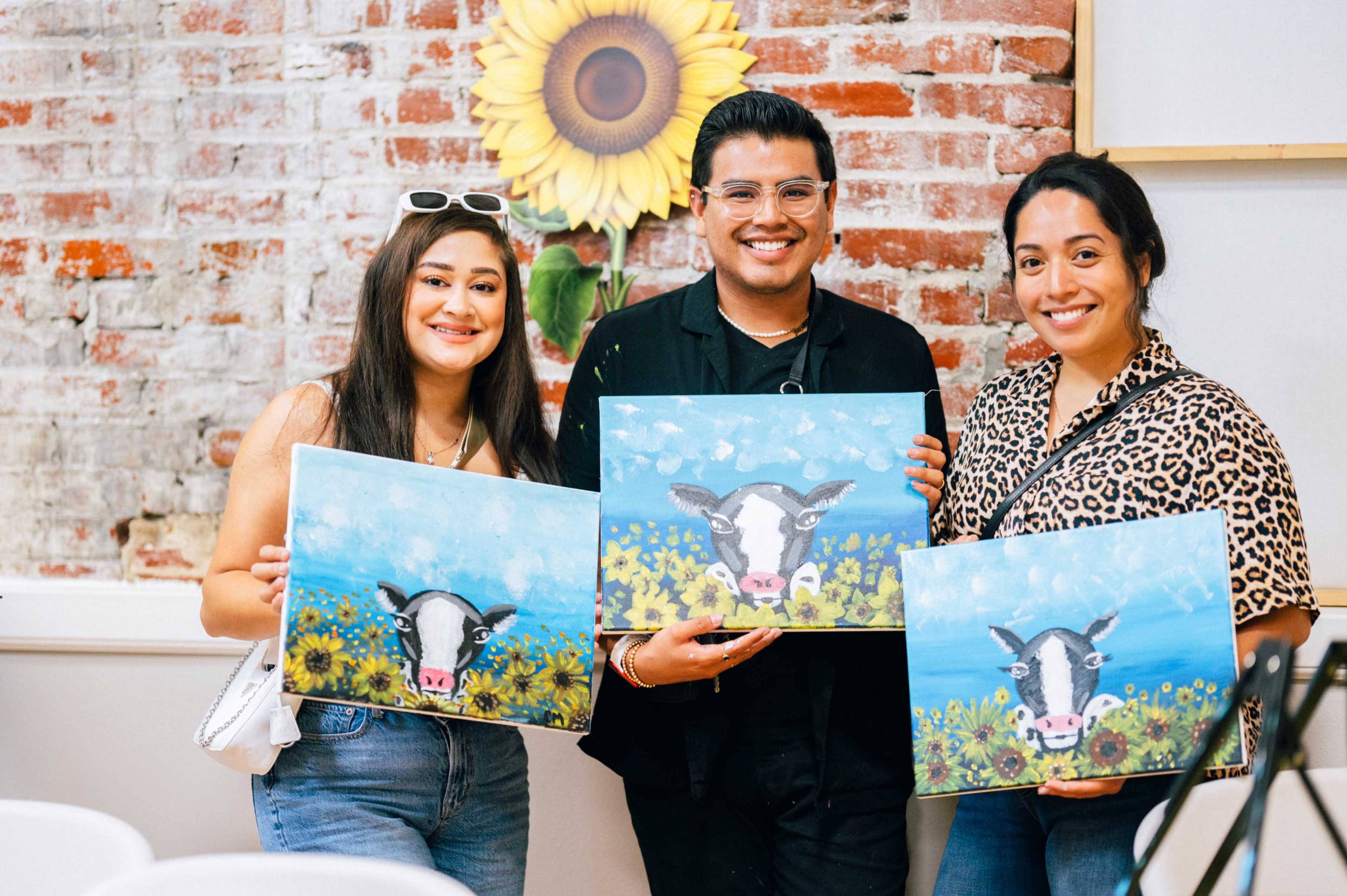 Three individuals hold paintings of a cow among flowers in a venue with a brick wall and a sunflower decoration.