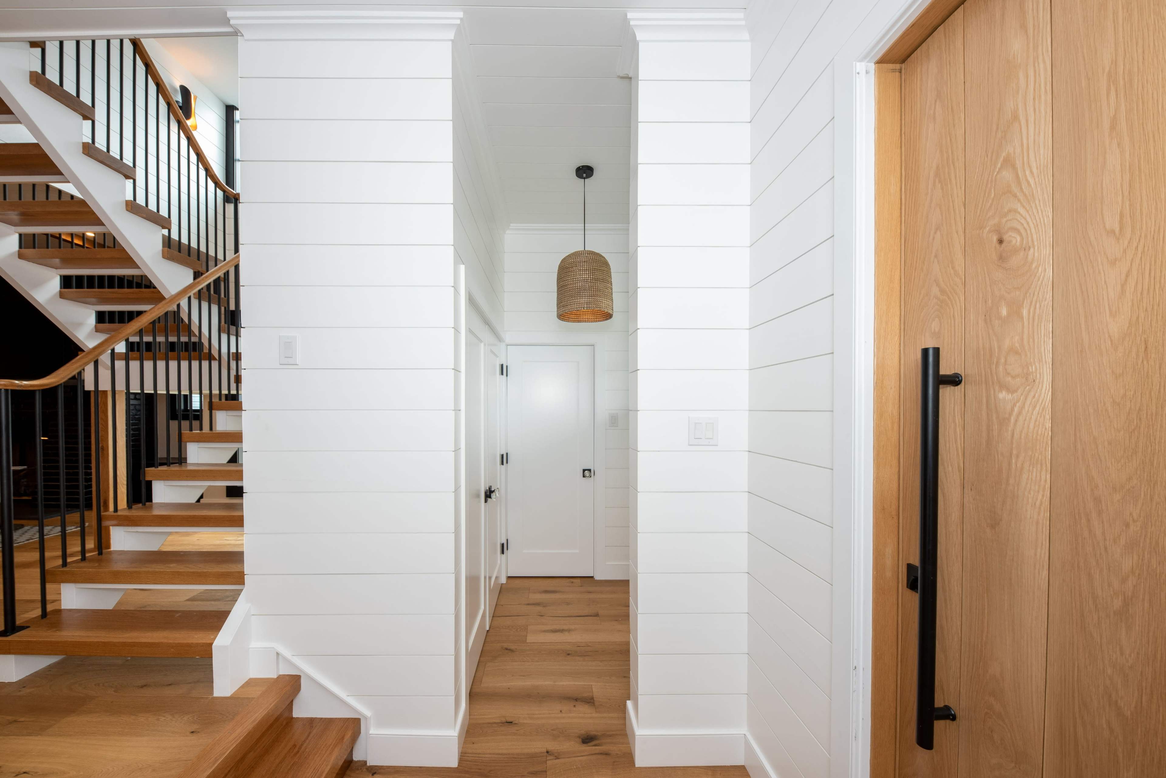 A bright entryway with white shiplap walls, wooden stairs on the left, and a light fixture hanging above a door on the right.