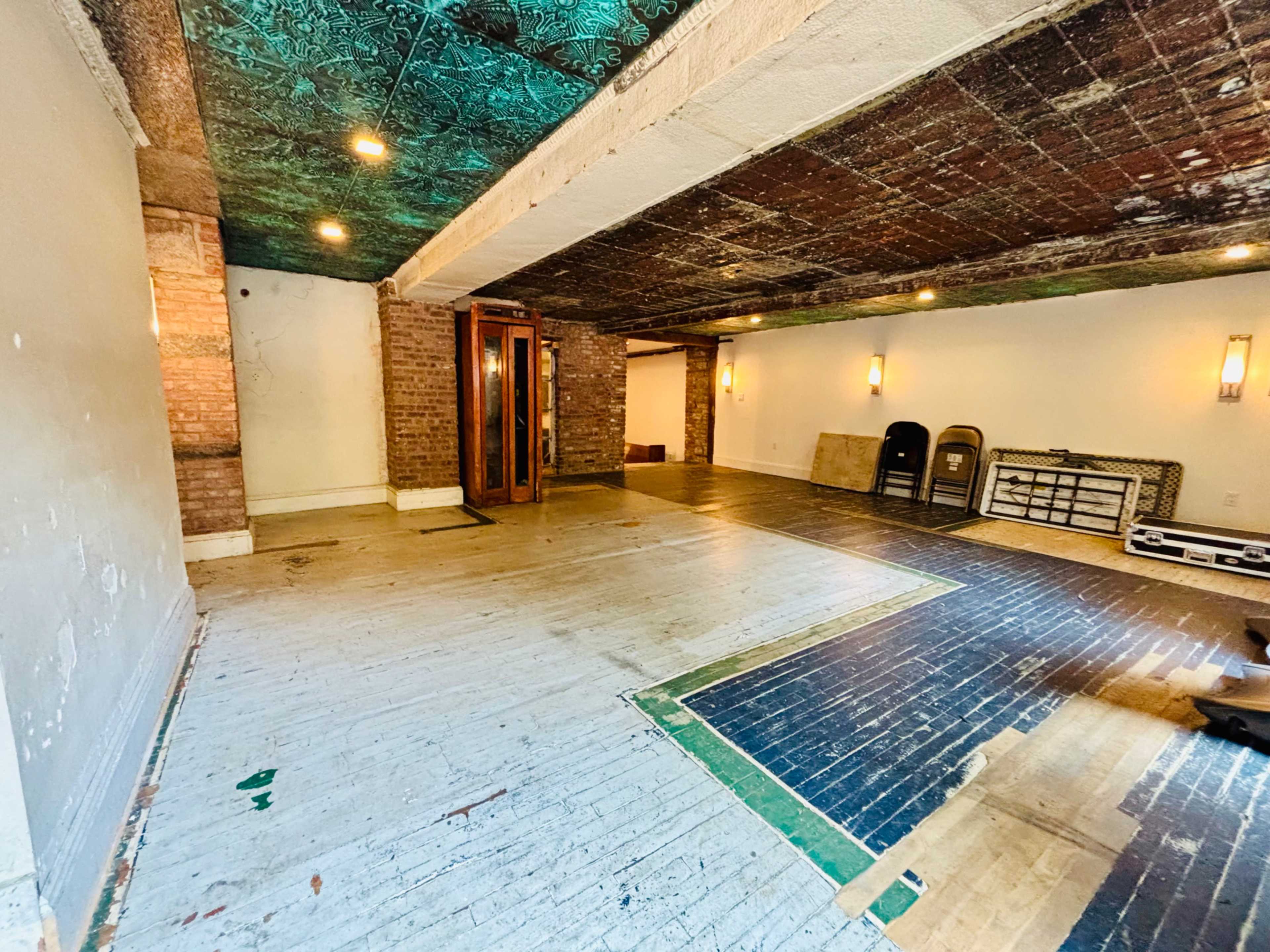 The image shows an empty room with exposed brick walls, a patterned ceiling, and various flooring materials, including wood and painted sections.